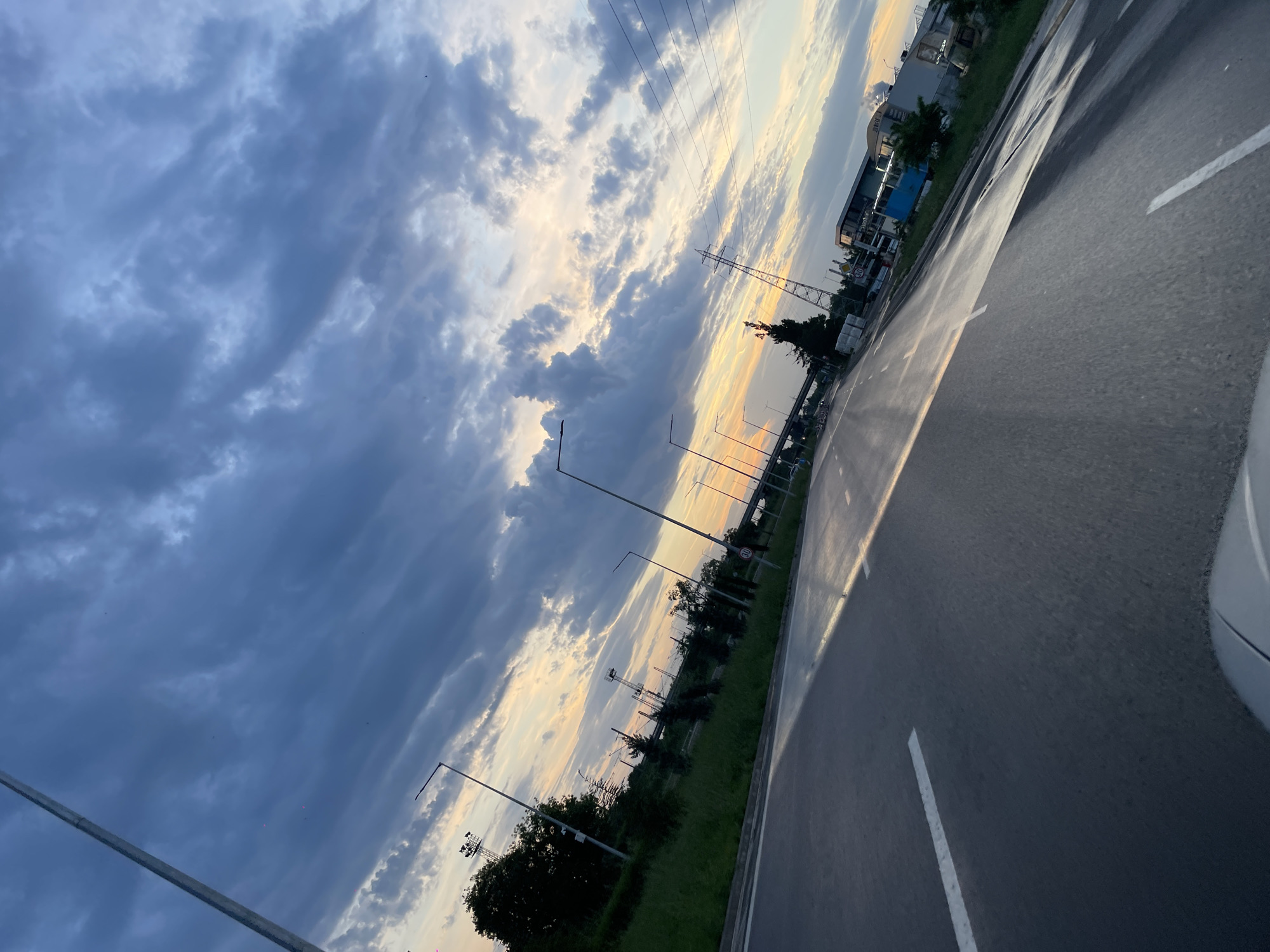 The image depicts a view from a vehicle on a road in Burgas, Bulgaria, likely at sunset or sunrise. The road is asphalt and marked with white lane dividers. Adjacent to the road is a grassy area with interspersed trees and lighting poles. Further in the background, a building is visible. Above, the sky is filled with layered clouds exhibiting shades of blue and orange. A power line is stretched across the sky.