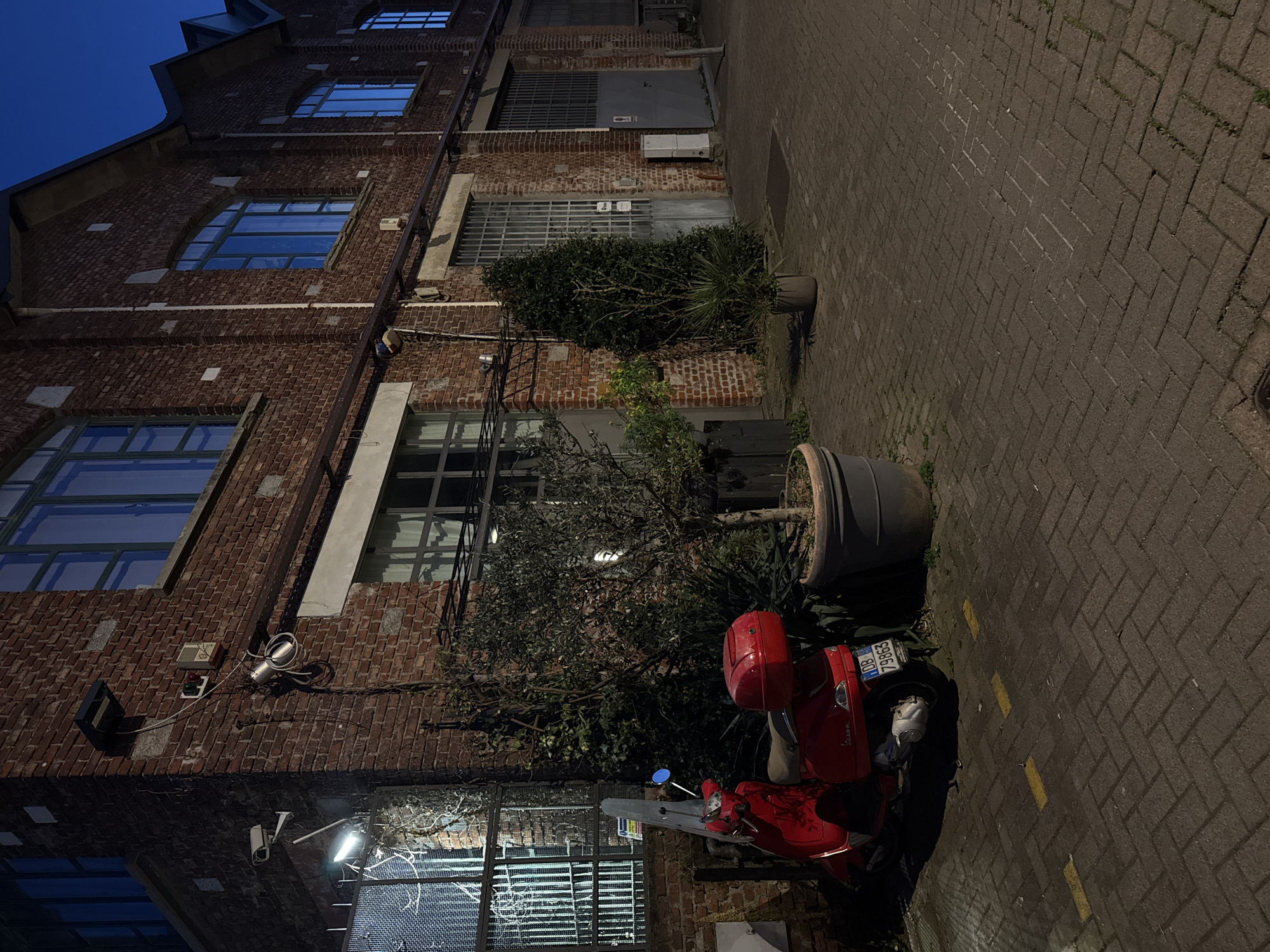 The image shows a street scene in Milan, Italy, at dusk. A red Vespa scooter is parked on a brick-paved sidewalk next to a large planter. The license plate is visible and reads "DB 79653." The scooter is partially obscured by a small tree or shrub. The sidewalk runs alongside a multi-story brick building with several windows, some with security bars. Various fixtures, including lights, security cameras, and conduits, are attached to the building's facade. The overall lighting is dim, with artificial lights illuminating portions of the building and sidewalk. The sky is a deep blue, indicating twilight. There are no people visible in the frame.