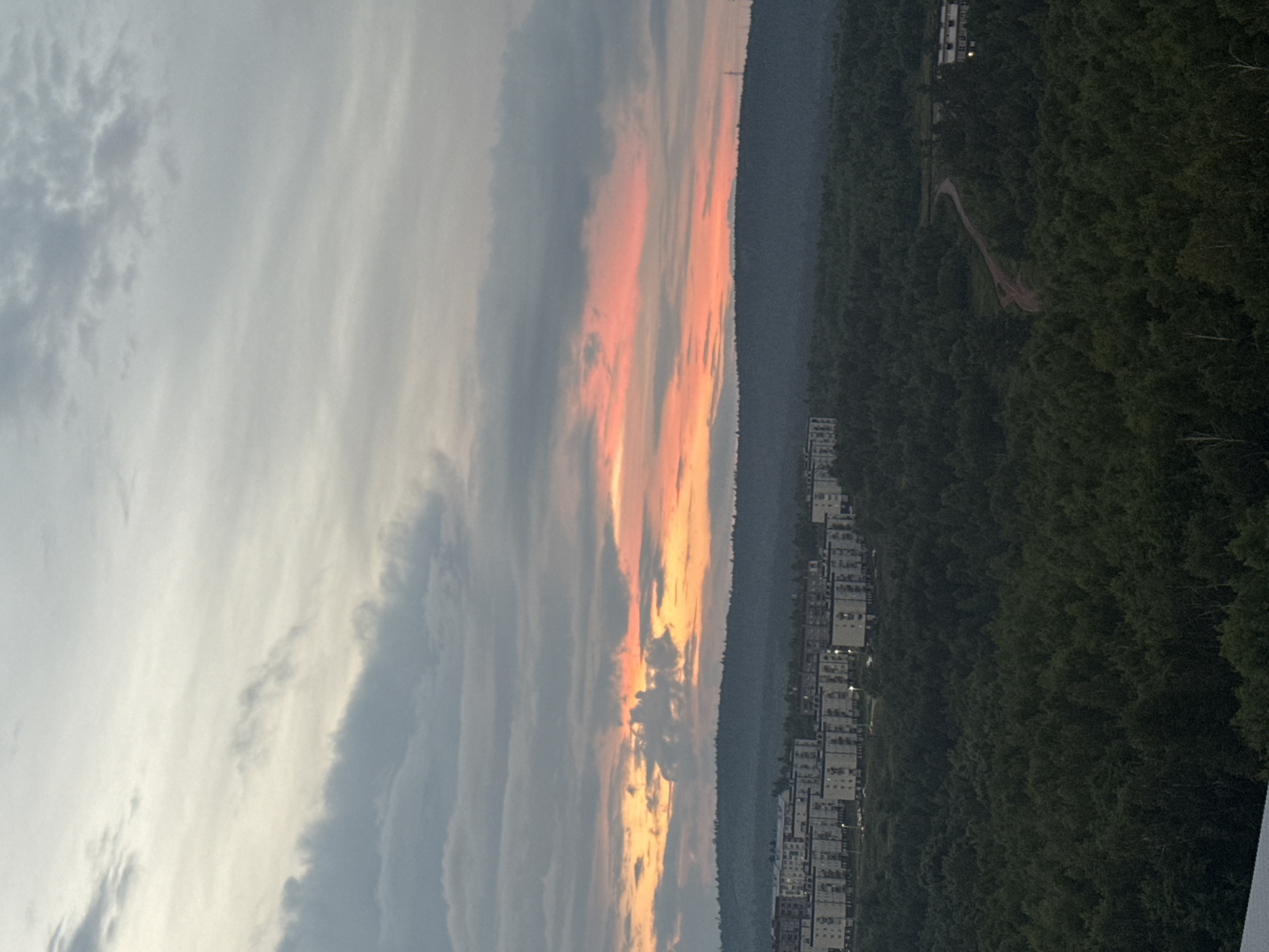 This media file captures a panoramic view of a cityscape nestled beside a dense forest, with a dramatic sunset painting the sky. The scene is oriented vertically, with the sky occupying the left half and the landscape the right.
The sky is filled with a breathtaking display of sunset colors. Wispy, elongated clouds stretch across the frame, transitioning from soft grays and whites to vibrant hues of orange and pink where the sun's rays pierce through. The lighting suggests it is either sunrise or sunset.
On the right side of the image, a dark, heavily wooded forest stretches along the horizon. The trees are a deep green, indicating a healthy, mature forest. Emerging from the dense foliage is a series of multi-story residential buildings, appearing as rows of white and light-colored structures. A winding dirt path or road is visible through the trees, adding a touch of human presence to the natural landscape. The presence of buildings suggests a settlement or town on the edge of the forest.
The overall mood of the image is serene and majestic, with the beauty of the natural sunset contrasting with the organized human development. The time of day is clearly indicated by the sunset. The location cues, such as the dense forest and the style of apartment buildings, combined with the provided location context of Bratsk, Russia, suggest this is a view from an elevated position overlooking a residential area adjacent to a forest in that region. There is no visible text in the image.