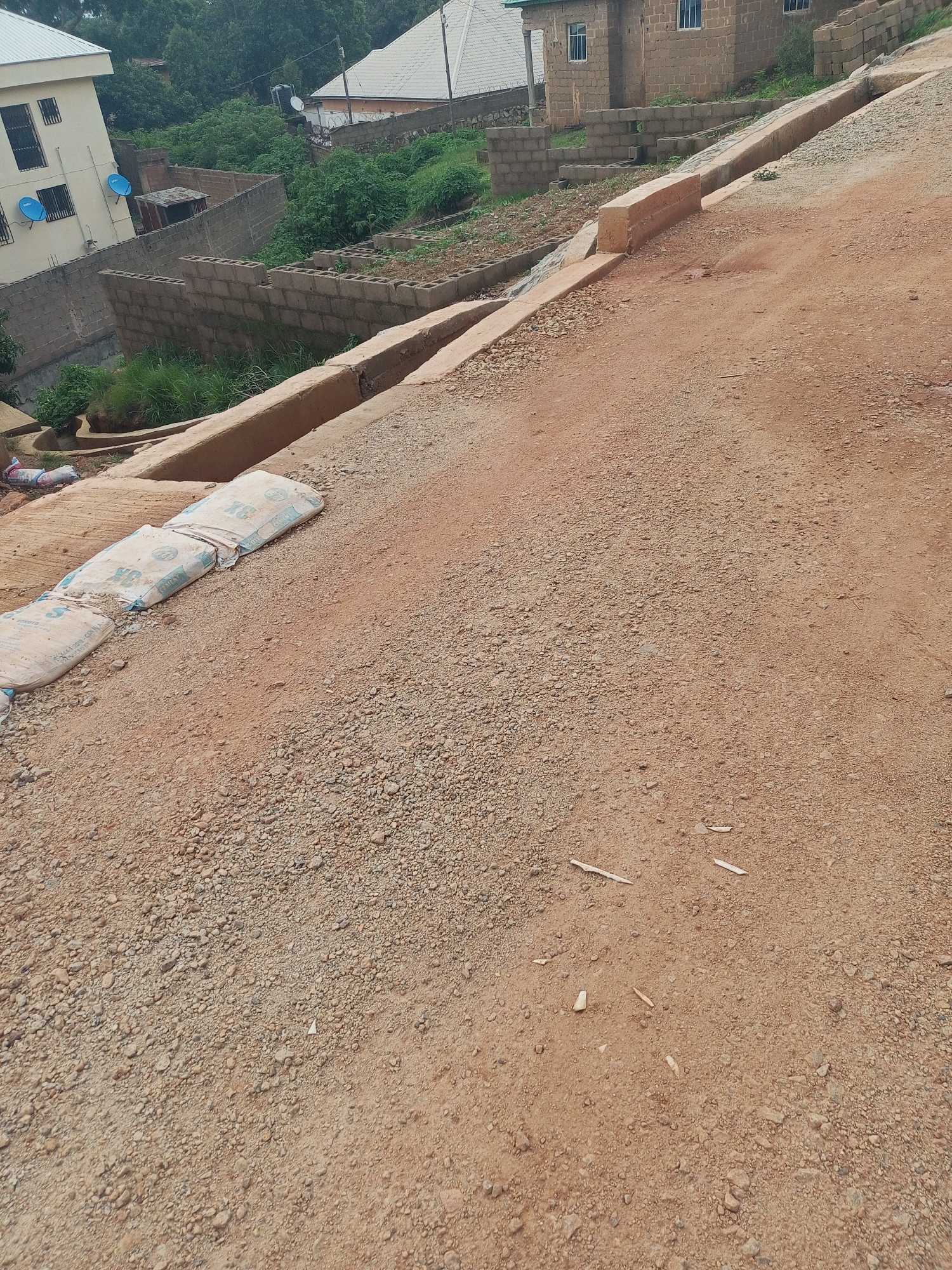 The image depicts a street scene in Jos, Nigeria. The foreground is dominated by a reddish-brown dirt and gravel road, with a concrete drainage channel running along its left side. Several bags, likely containing cement, are placed on the edge of the drainage channel. In the mid-ground, there are partially constructed brick walls, overgrown vegetation, and a few small buildings. One building appears to be a multi-story residential structure with blue satellite dishes mounted on its exterior. Further in the background, more residential buildings with tiled roofs are visible, along with dense green trees. The overall impression is one of ongoing construction and development in a semi-urban environment.