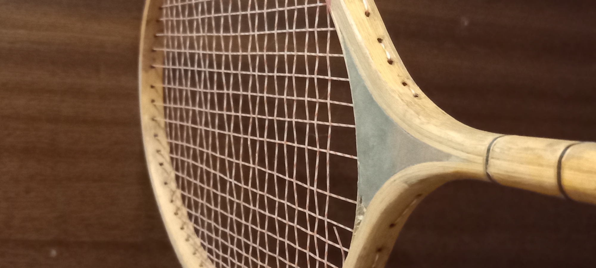 The image shows a close-up of a vintage wooden tennis racket. The racket is positioned at an angle, with the strings and a portion of the frame filling the majority of the frame. The strings are tightly woven and appear to be light pink or peach in color. The wooden frame is a light natural wood tone, with visible grain. A light blue or grey accent piece is visible at the throat of the racket, where the frame splits to form the handle. The background is a dark, textured brown, likely a wall or fabric, which is out of focus. There are no people present in the image, and no indication of the location within the city of Kryvyi Rih, Ukraine. The image is a still photograph.