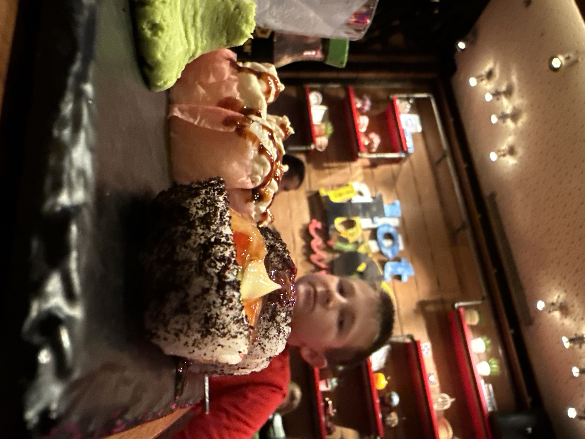 A child in a red shirt is featured holding a piece of sushi on a black tray. The sushi piece is covered in black sesame seeds and contains a white sauce and other ingredients. The tray also holds wasabi and other sushi rolls. The location appears to be a restaurant with shelves displaying various items. The child is looking toward the viewer, while the shelves in the background and the lights on the wall suggest the interior of an establishment in Sofia, Bulgaria.