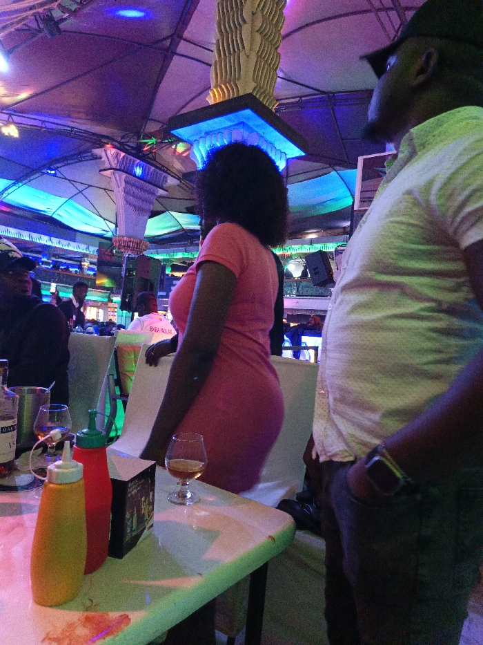 In a vibrant, colorfully lit indoor space, a woman in a pink dress stands facing away from the camera. A man in a white shirt and hat stands beside her, facing right. On a nearby table, there are bottles of mustard and ketchup, a glass of alcohol, and a small box. The background reveals other people seated and standing in the brightly decorated environment.