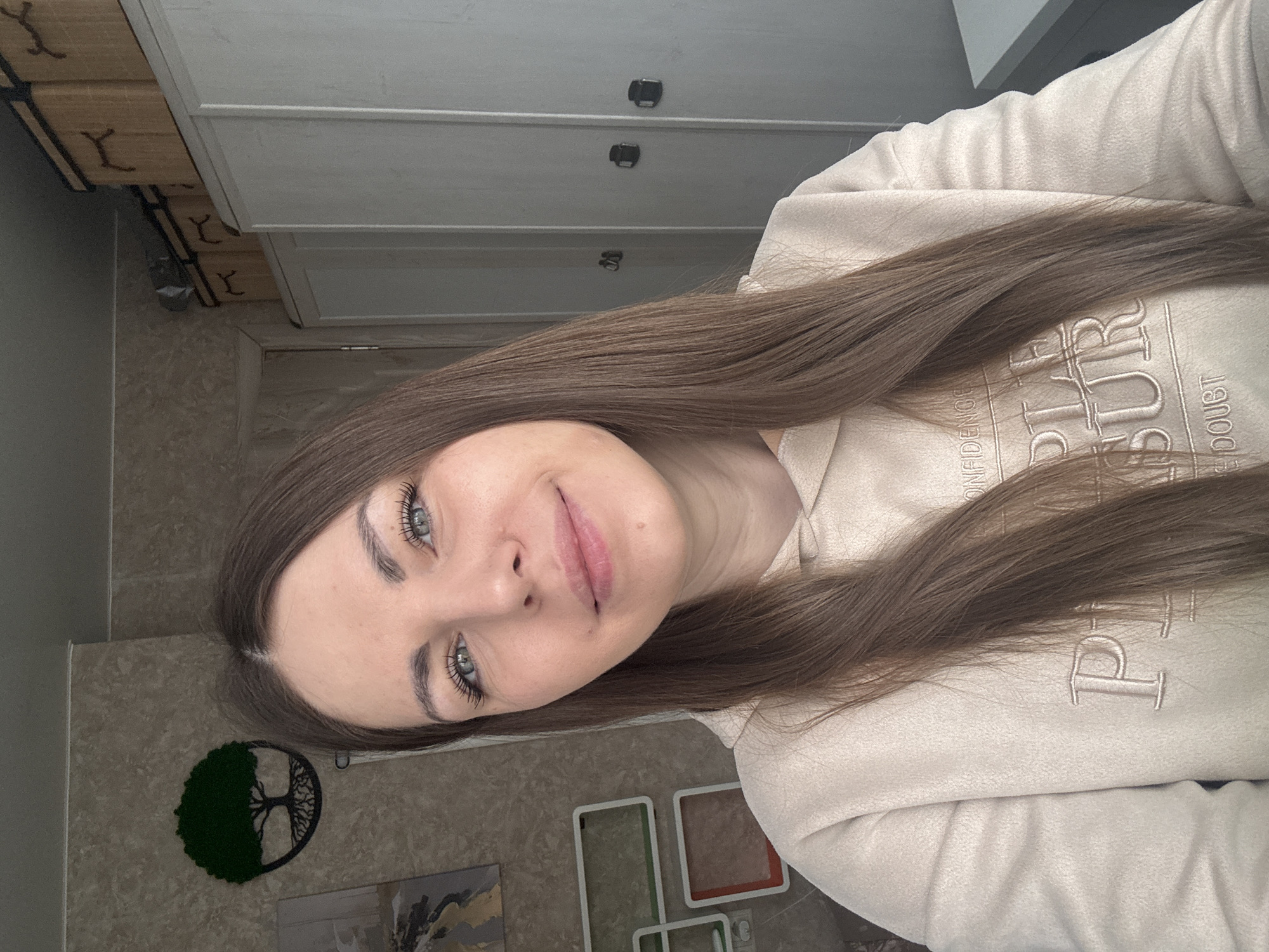 A young woman is captured in a selfie, taken indoors from a slightly angled perspective. She has long, straight, medium-brown hair, and light blue or grey eyes, enhanced with dark mascara. Her dark, well-defined eyebrows frame her face, which holds a subtle, natural smile, conveying a calm and pleasant demeanor. She is dressed in a light beige or cream-colored hooded sweatshirt.
The background reveals a domestic setting. To the upper left, a light-toned wooden dresser or cabinet with dark handles is visible, alongside what appears to be wooden crates and metal hooks on a lower shelf. The wall behind her is textured and decorated with a circular "Tree of Life" art piece, half-filled with green, moss-like material, and a framed abstract artwork in black, white, and possibly gold/yellow hues. Below these, several empty rectangular frames of varying sizes, some with colored accents, are arranged on the wall, suggesting a personalized living space. A small electrical outlet is also visible.
Embroidered text on her hoodie is partially discernable, with visible parts reading "CONFIDENCE," "PURE," and "NO DOUBT." The indoor lighting suggests it is daytime. The scene depicts a casual moment in a home environment located in Раздолье, Russia, with the woman posing directly for the camera.
