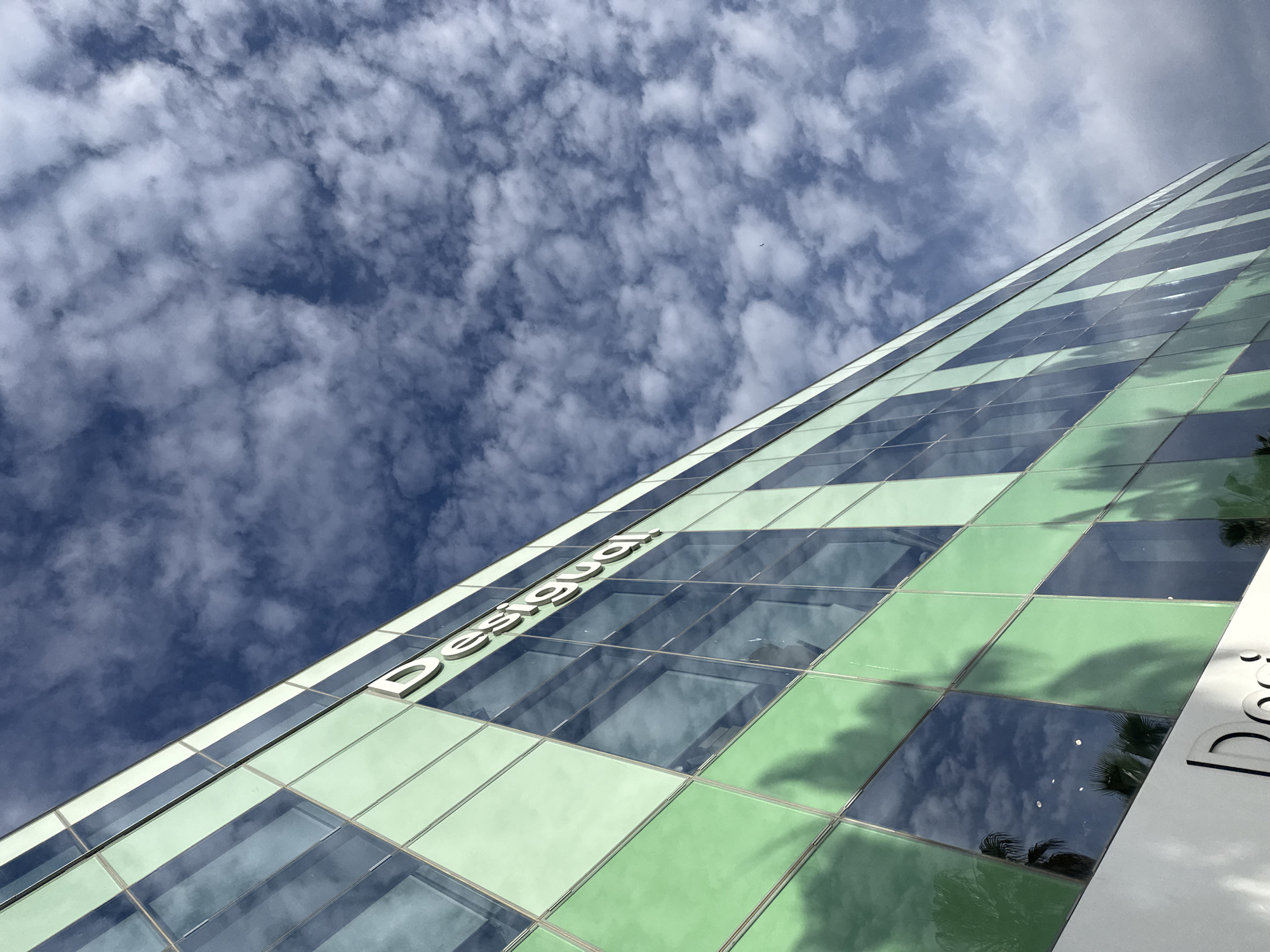 The image presents a low-angle perspective of a building against a sky filled with scattered clouds. The building's facade is characterized by a grid-like pattern of alternating green and blue tinted glass panels, bearing the word "Desigual" in large white lettering. The glass panels reflect the sky, mirroring the cloud formations. The scene's location is indicated by the signage to be Barcelona, Spain.