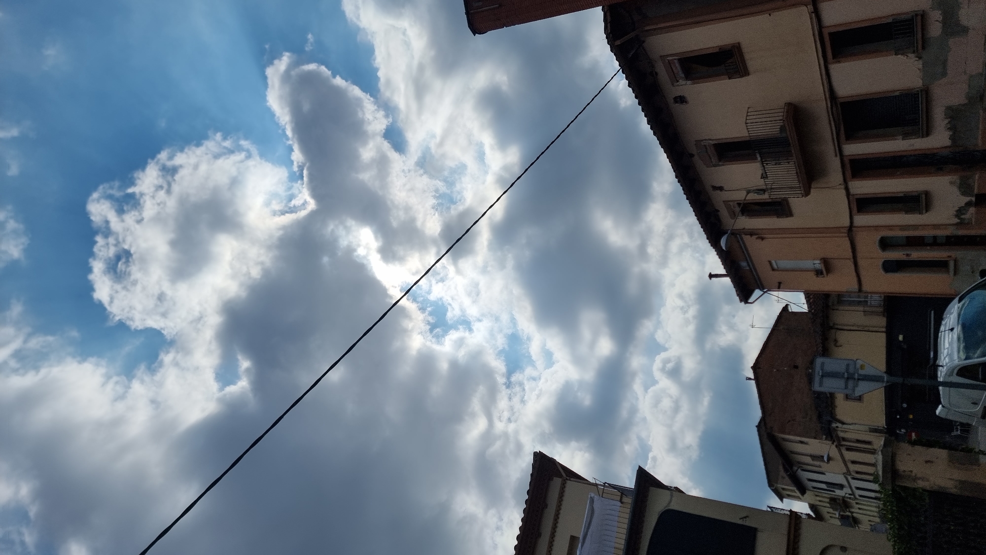 The image depicts an upward view of buildings and the sky, with a wire bisecting the frame diagonally from the upper left to the lower right. The sky is filled with dramatic, cloudy formations against a blue backdrop, with sunlight filtering through some of the clouds, creating bright patches and rays.
In the foreground and midground, parts of buildings are visible. On the left, a portion of a building with a tiled roof and a balcony is seen. Further to the right, other buildings with tiled roofs and windows, some with balconies, are present. The architecture suggests a European urban setting. One building is painted in pale orange, another in a lighter cream color. A small white van is partially visible at the bottom center of the image. A traffic sign on a pole is also visible in the lower middle section.
The scene is set in a street in the city of Olot, Spain, characterized by its urban architecture and the presence of residential or commercial buildings. The weather appears dynamic, with a mix of sun and clouds. No people are directly visible in the frame.