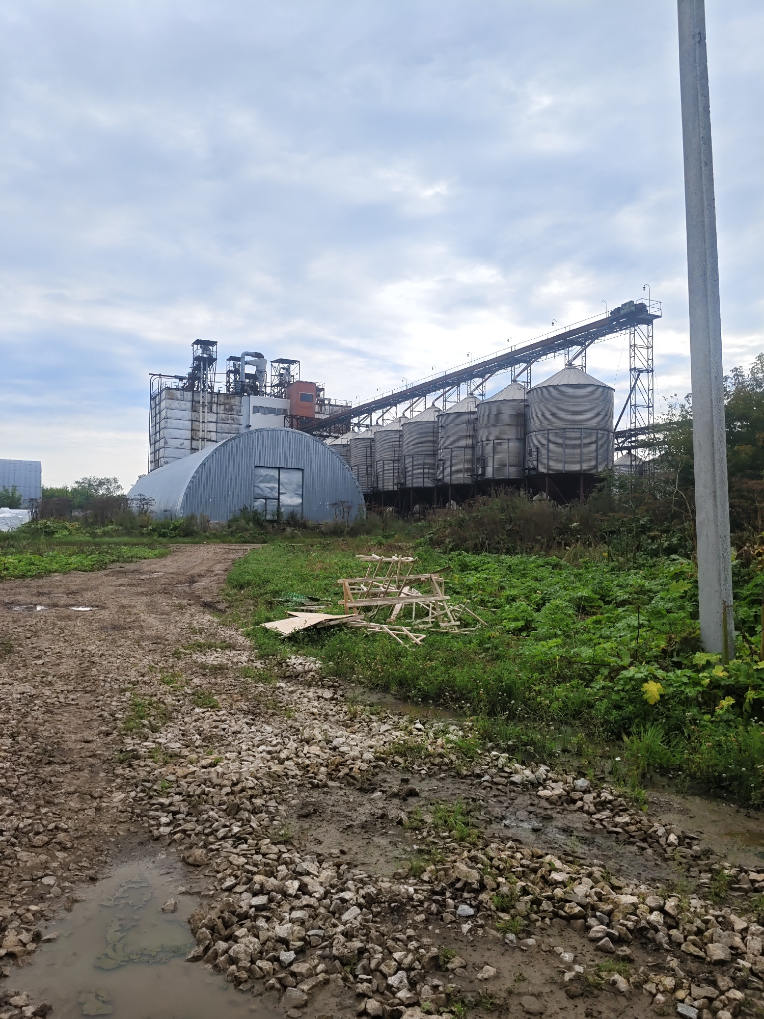 This is a photograph of an industrial facility in Platoschino, Russia, under a cloudy sky. In the background, a large structure with multiple cylindrical silos and a conveyor belt system dominates the scene. To the left of the silos, a long, arched metal building with a large window is visible. The foreground consists of a dirt path littered with rocks and a muddy puddle. Dense green vegetation surrounds the industrial buildings, with trees to the far right. A concrete utility pole stands tall on the right side of the image. There are no people or animals present in the photograph.