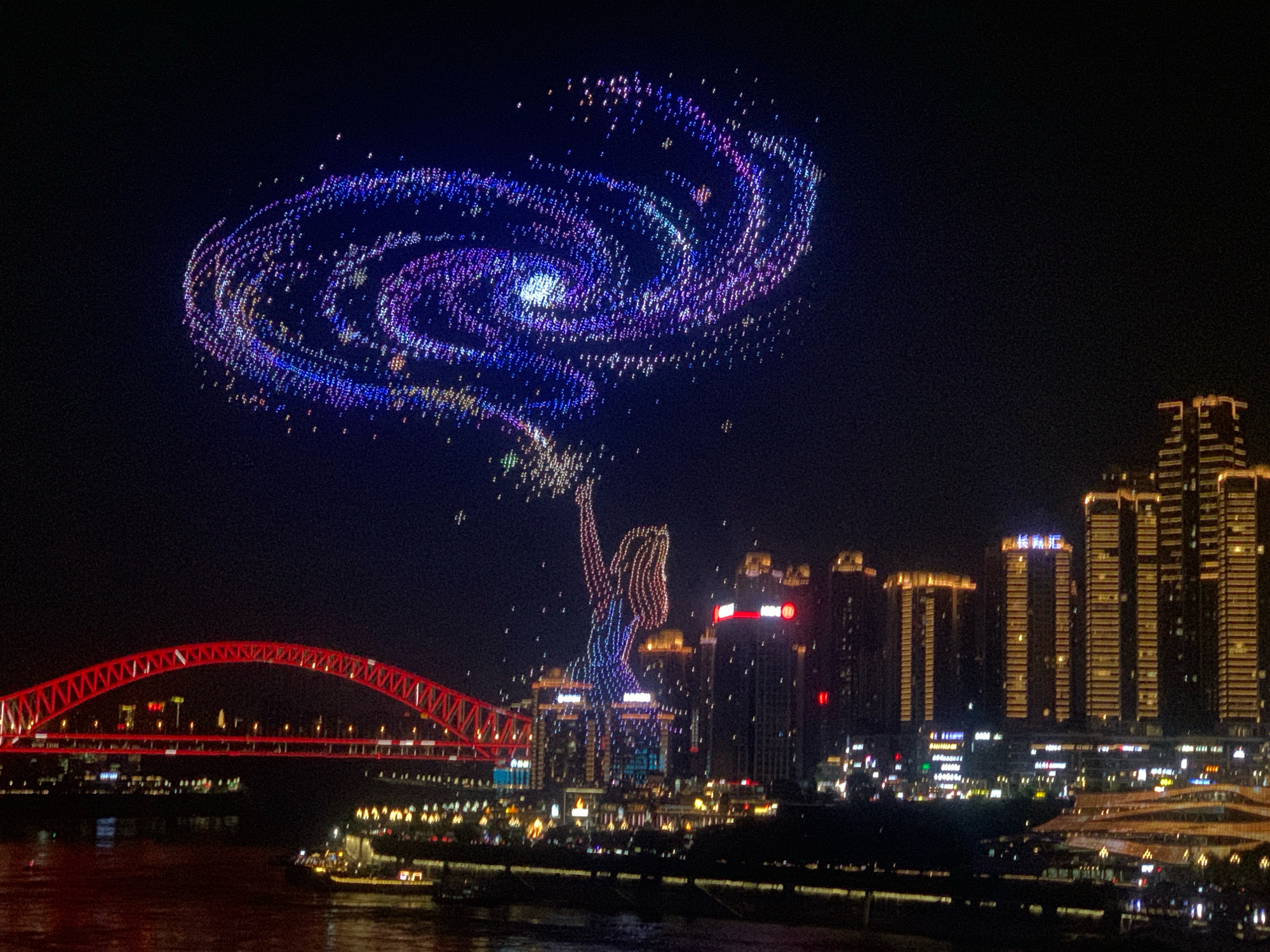 A drone light show is taking place at night over a city skyline in Yuzhong District, China. A large spiral galaxy formation, composed of numerous small lights in shades of blue, purple, and white, is visible in the upper portion of the sky. Below the galaxy formation, a human-like figure, also made of lights, is depicted with its arm raised, appearing to release the galaxy into the sky. The figure is illuminated in shades of blue and purple. In the foreground, a red illuminated bridge spans a body of water. On the right side of the image, tall buildings with illuminated windows form a city skyline. Several boats are visible on the water. The scene captures a nighttime urban landscape with a spectacular aerial display.