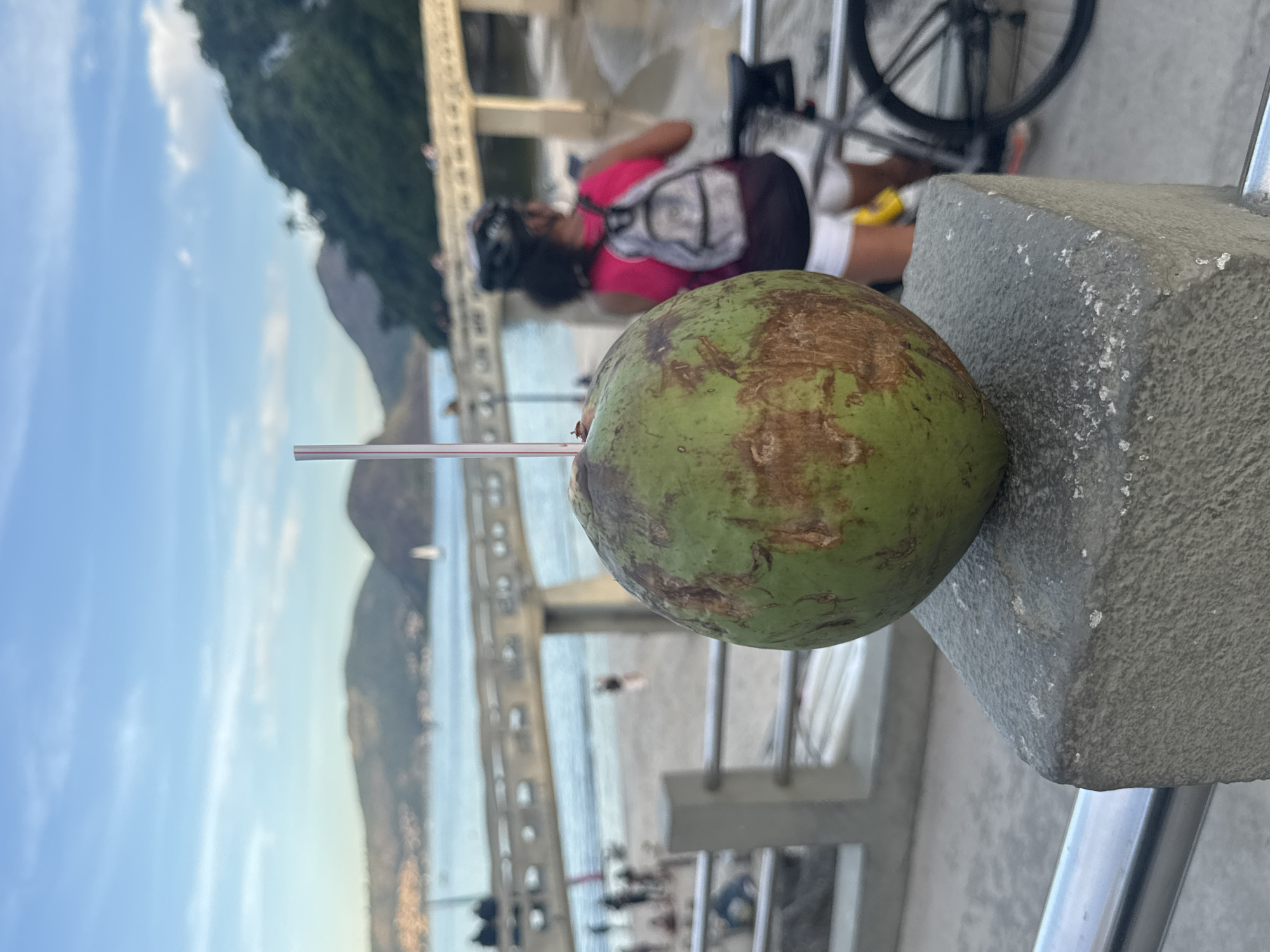 The image depicts a close-up of a green coconut with a straw, placed on a stone pillar. In the background, the scene transitions to a blurry view of the Icaraí beach and the Ponte Rio-Niterói bridge in Niterói, Brazil. The sky is a pale blue with some clouds. A woman wearing a helmet, pink top, and white shorts is visible on a bicycle, facing away from the camera. Other people and a sailboat are present on the beach and in the water, respectively. The overall ambiance suggests a relaxed, outdoor setting in a coastal city.