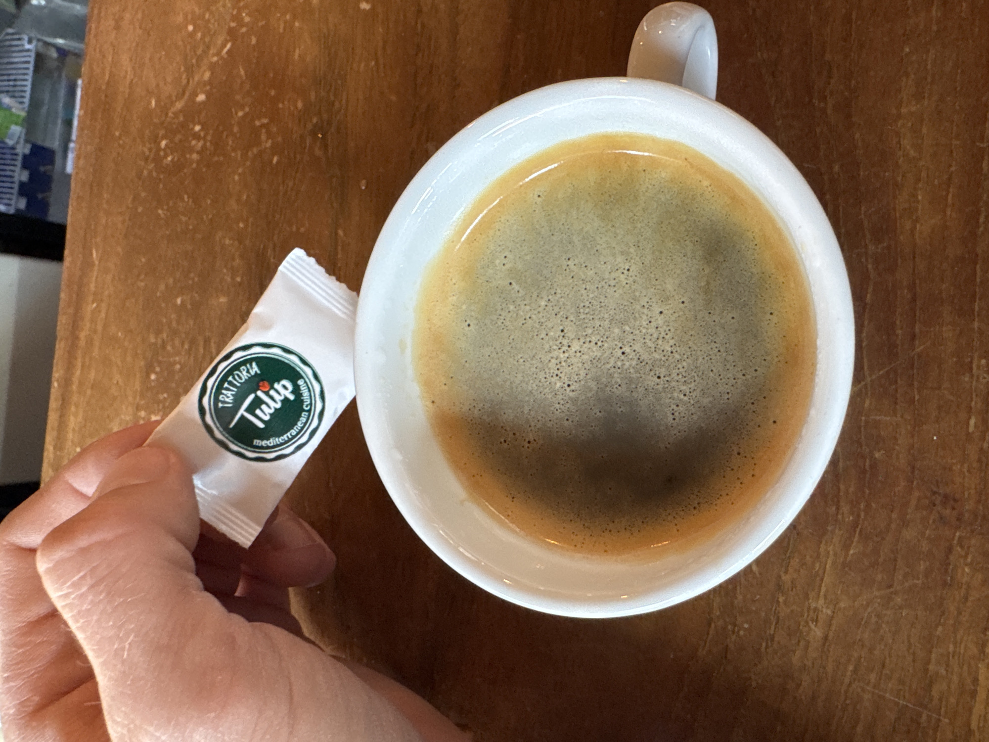 The image, taken in Rafailovići, Montenegro, presents an overhead view of a hand holding a white sugar packet next to a white ceramic coffee cup, all resting on a wooden table. The cup is filled with dark coffee, topped with a visible layer of frothy crema, suggesting an espresso or similar coffee drink. A small white handle extends from the upper left side of the cup.
A human hand, with visible skin lines and a light complexion, holds the white, rectangular sugar packet. The packet features a circular green logo. Text on the logo reads "TRATTORIA" arched over the word "Tulip" in a stylized script, followed by "mediterranean cuisine" in a smaller font at the bottom.
The background consists primarily of a warm-toned wooden surface, likely a table, displaying its natural grain. In the upper left corner, out-of-focus elements, possibly part of a counter or shelves, are dimly visible, suggesting an indoor setting like a cafe or restaurant. The scene captures a quiet moment, possibly during a coffee break.