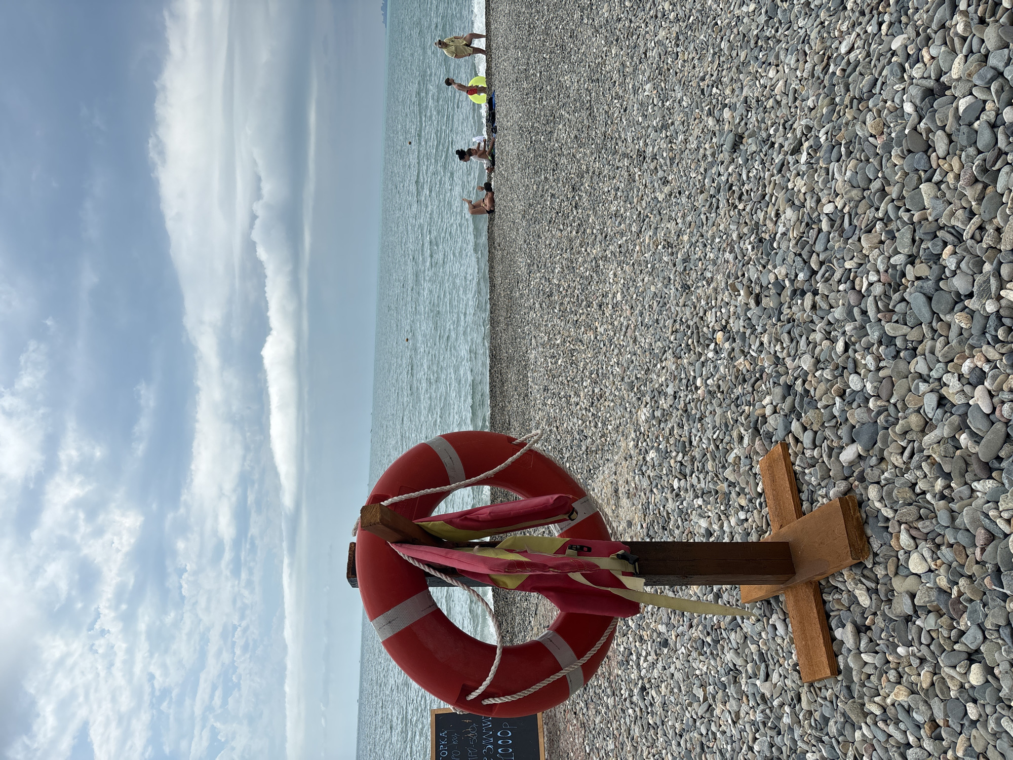 On a pebble beach in Sochi, Russia, a red life preserver is displayed on a wooden stand. Two child-sized life jackets, one pink and one yellow, are hung on the life preserver. A chalkboard sign to the left of the life preserver displays text in Russian, including prices. In the background, the sea stretches to the horizon under a cloudy sky. Several people are visible on the beach near the water, including a person lying on their back, two adults and two children sitting, and a woman standing.