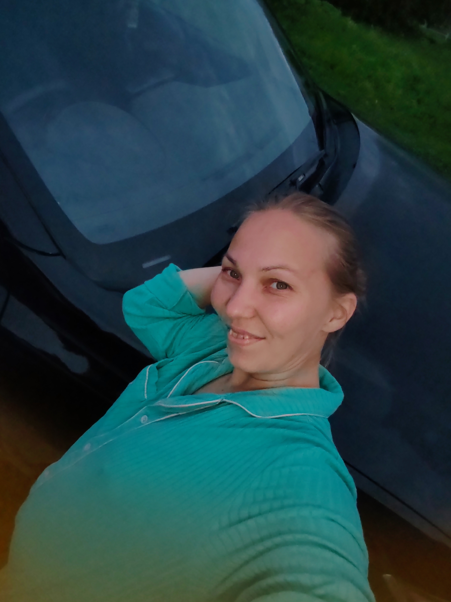A medium-shot, eye-level, selfie-style photograph shows a blonde woman with light skin, wearing a teal pajama top with white trim and a button. She is positioned with her right arm behind her head, looking towards the camera with a smile. The background is dark, with the reflection of something round and light appearing on the windshield of a car, and a strip of green grass visible at the top right. The image appears to have been taken outdoors at dusk or in low light conditions, potentially in or near the city of Chuchloma, Russia, though the specific location is not confirmed by visual evidence.