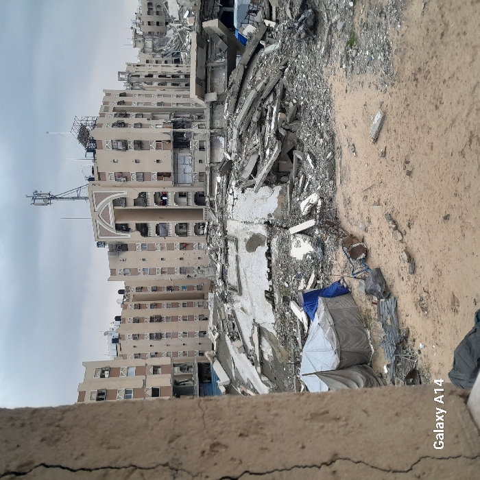 The image portrays an urban scene in Khan Yunis, Palestinian Territory, dominated by widespread structural destruction under an overcast sky. In the immediate foreground on the left, a cracked, light-colored concrete pillar is partially visible, bearing "Galaxy A14" text at its lower edge. The ground transitions from disturbed sand with scattered debris to an extensive field of rubble in the midground. This central area is a dense accumulation of broken concrete slabs, twisted rebar, and pulverized building materials, indicative of collapsed structures. Amidst the debris, a segment of a white concrete dome or arch is discernible, and two light-colored canvas tents, one partially covered by a blue tarp, are pitched on the sandy ground to the left of the main rubble pile. In the background, multiple multi-story buildings, predominantly beige with distinctive arched decorative elements and balconies with white railings, stand beyond the devastation. Many of these background structures exhibit significant damage, including numerous small holes or impact marks on their facades and broken windows. A prominent telecommunications tower with multiple antennas is mounted on the roof of the central background building. No individuals are visible in the scene.