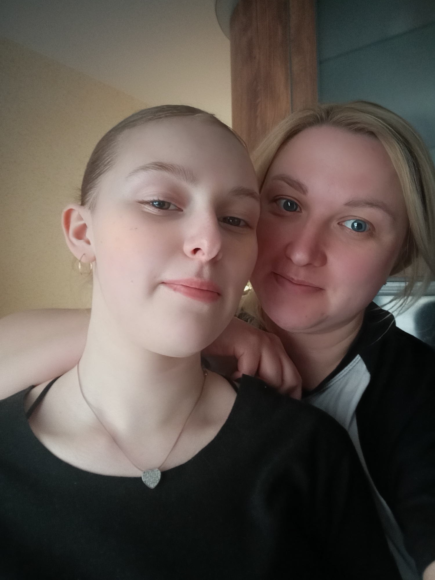 A close-up shot shows two women posing for a selfie. The woman on the left is younger, with light skin, light brown hair pulled back, and a small gold hoop earring on her left ear. She wears a black top and a delicate necklace with a heart-shaped pendant. The woman on the right has blonde hair styled around her face and blue eyes. She wears a black and white jacket. Her arm is around the younger woman's shoulder. The background appears to be indoors, with a light yellow wall on the left and a wooden structure behind the women. It is unknown if the location is Chaikovsky, Russia.