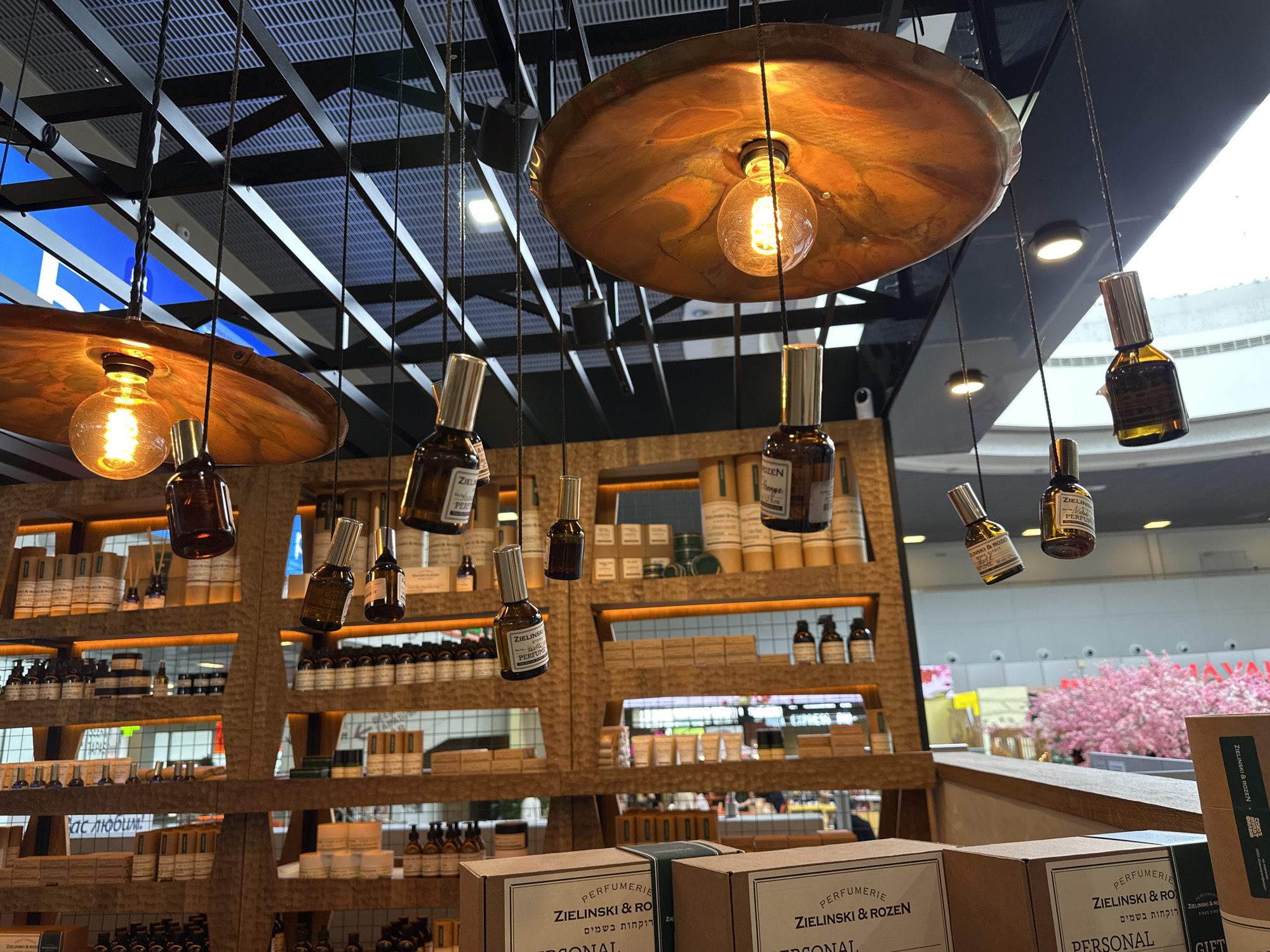 The image shows the interior of a Zielinski & Rozen perfumery, possibly located at Pulkovo Airport in Saint Petersburg, Russia. The foreground features several boxed Zielinski & Rozen products with Hebrew script. The middle ground showcases a wooden shelving unit stocked with various perfumes, lotions, and other cosmetic items. Above, decorative light fixtures with Edison-style bulbs hang from the ceiling, with perfume bottles suspended on strings from the fixtures. In the background, out of the storefront, a glimpse of the airport terminal is visible, featuring a cherry blossom tree and terminal signage.