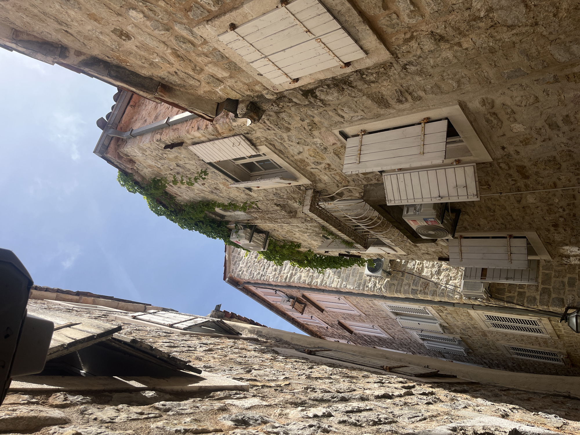 The image shows a view upwards between two buildings in an alleyway in Unknown, Unknown. The buildings are constructed of stone. One building has several windows with white wooden shutters, some open. There are air conditioning units attached to the side of the building. Green foliage grows on the side of one building. The sky is visible at the top of the image.