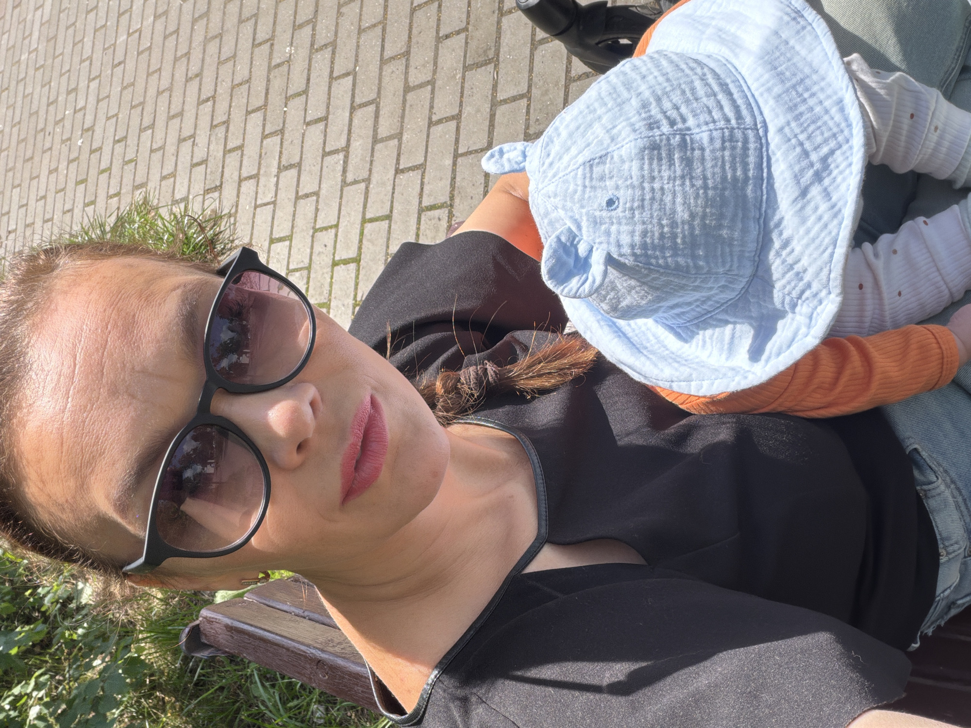 A woman with sunglasses and pink lipstick is taking a selfie with a baby. The woman has her hair in a braid and is wearing a black shirt and blue jeans. The baby is wearing a blue hat with ears and an orange shirt. They are sitting on a bench in a park with a brick pathway behind them. The image is taken from a high angle.