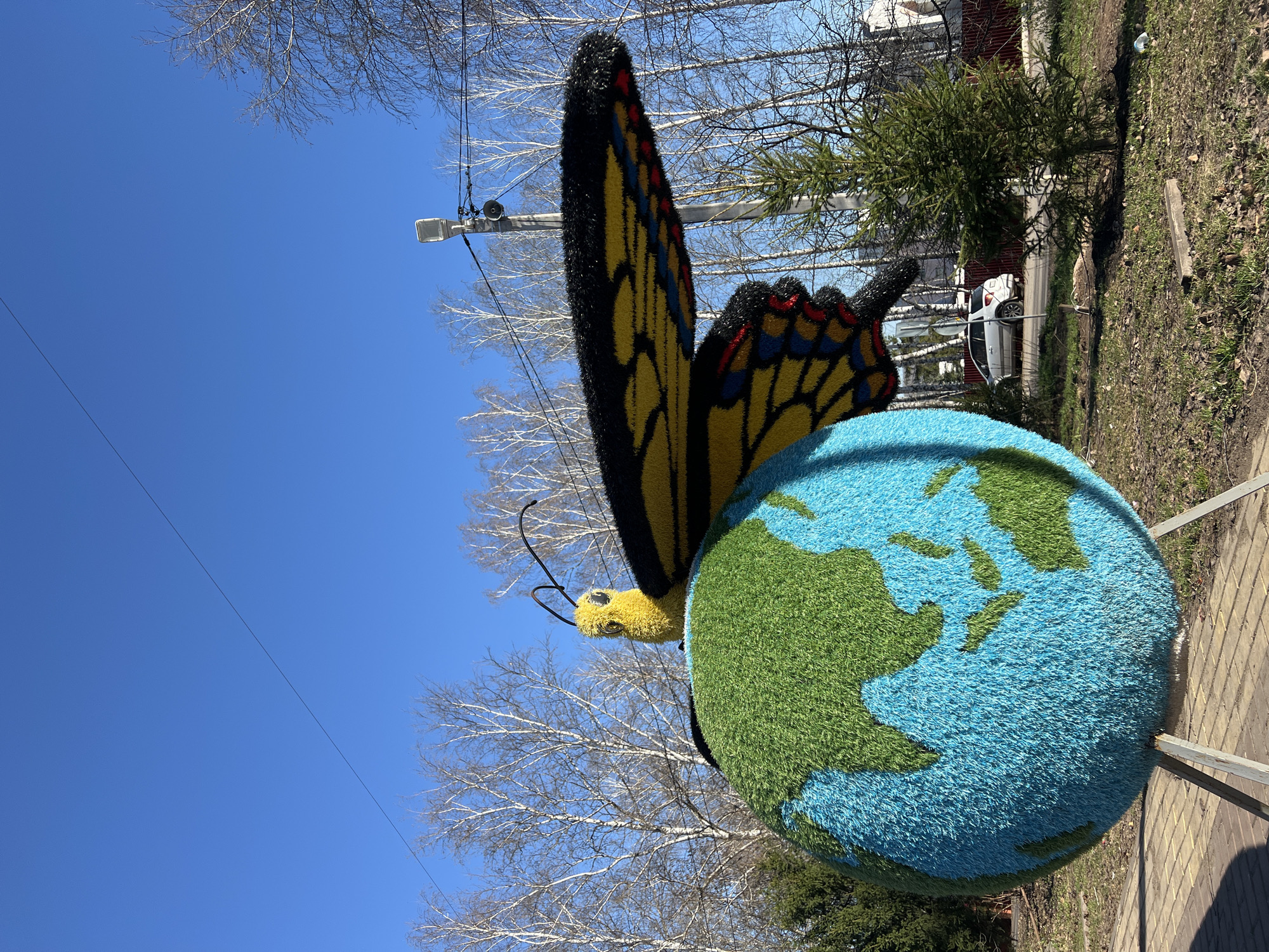 An outdoor scene in санатория Юматово имени 15-летия БАССР, Russia, features a decorative display. A large, spherical representation of the Earth, covered in blue and green fiber, rests on a stand. A large butterfly figure, constructed from similar materials in black, yellow, and red, is positioned on the globe. The blue sky is visible in the background, along with bare trees, a streetlight, and a glimpse of a car parked on a street. A green coniferous tree and a patch of brown earth are also in the background. The display sits on a paved area.