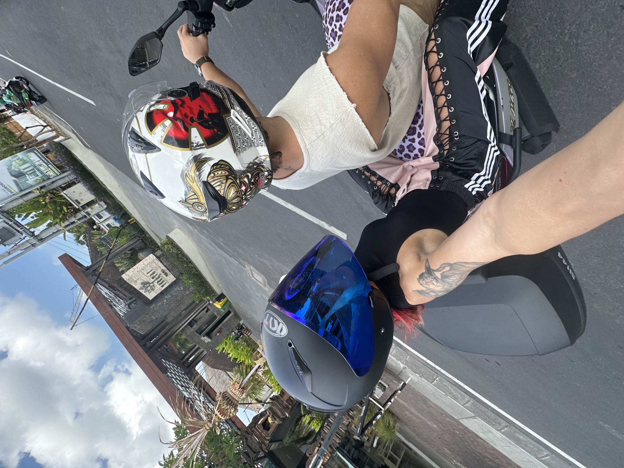 In North Kuta, Indonesia, two individuals are riding a scooter on an asphalt road. The person in the foreground, facing away from the camera, wears a black helmet with a blue reflective visor and a black top, with bright pink hair visible. Their arm, bearing a tattoo of a bird, is extended towards the viewer, holding a scooter handlebar. The person in the background, also facing away, wears a white helmet with a graphic design of a samurai warrior and a ripped beige t-shirt. They are seated behind the first individual. The scooter is a modern model with black storage attached to the rear. The environment features a modern building entrance with wooden beams and stone accents, a sign with a lotus flower symbol, and lush green foliage. The sky is blue with scattered white clouds. In the distance, other vehicles are visible on the road.