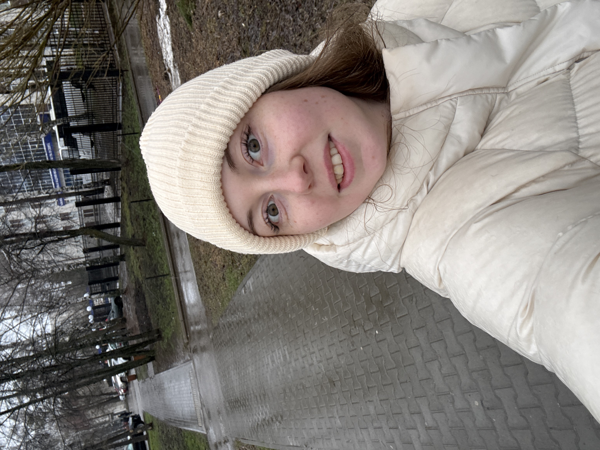 A young person is featured in a close-up selfie, looking directly at the camera with a gentle smile. They have fair skin with freckles and light-colored eyes, framed by light brown hair. They are dressed for cool weather in a cream or off-white knitted beanie and a matching puffer jacket.
The background shows an outdoor urban or suburban setting. The person appears to be on a wet, paved pathway made of interlocking grey concrete pavers, indicating recent precipitation. To their side, there is a narrow strip of damp ground with sparse grass and bare trees, suggesting late autumn, winter, or early spring. Beyond the trees, a dark metal fence separates the path from a street where several cars are parked alongside lampposts. In the distance, a multi-story building with dark windows and a faint blue sign (unreadable) is visible. The overall lighting is soft and diffused, consistent with an overcast day. No discernible text is visible within the image.
