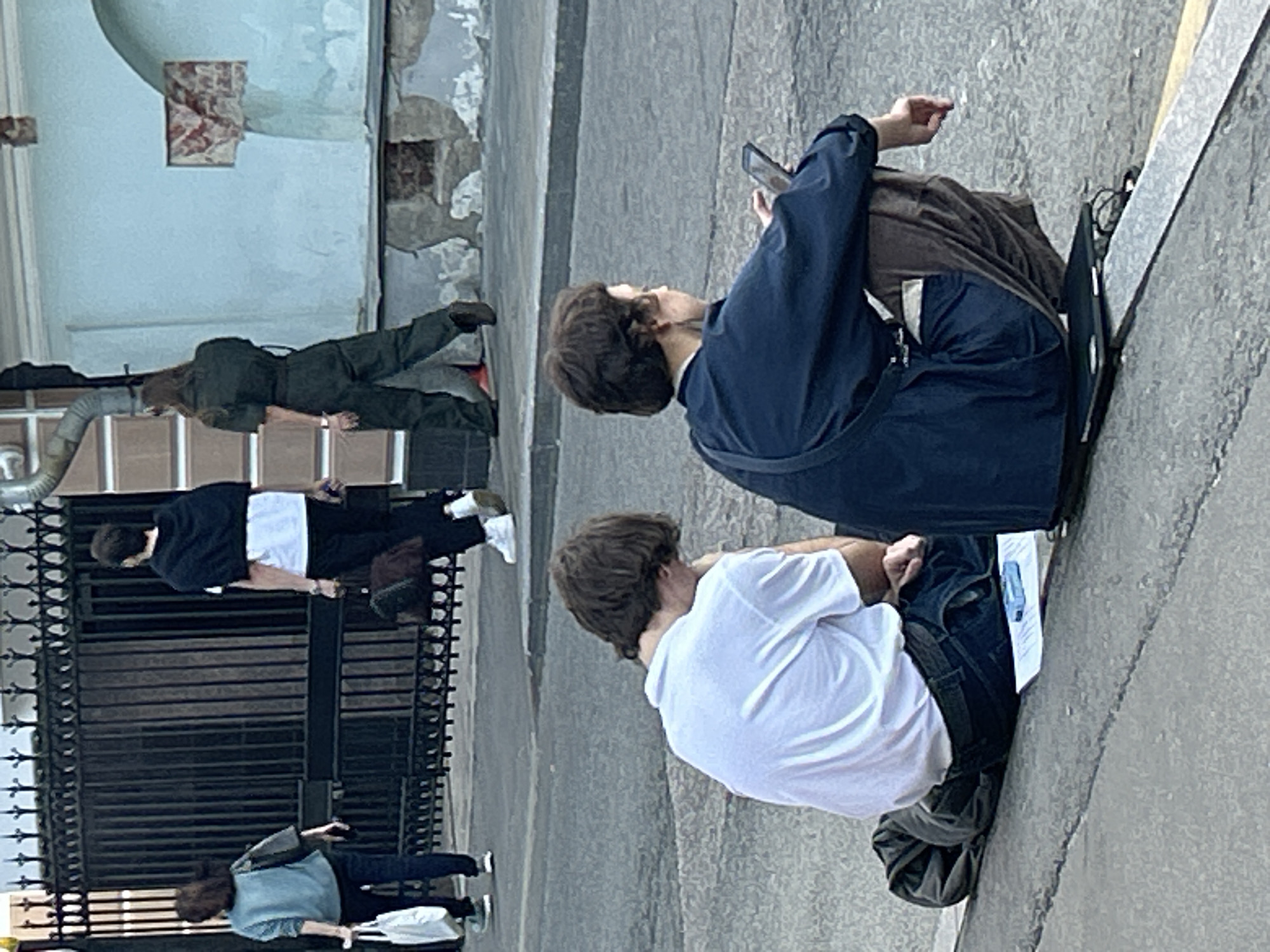 The image shows a street scene in Unknown, Unknown. Two people are sitting on the curb, one wearing a blue jacket and brown pants, and holding a mobile phone in hand. The other person wears a white shirt. Another person, wearing a green jumpsuit, is lying on a ledge of a building in the background. A black iron gate sits on the left. Two people walking can also be seen in the background.