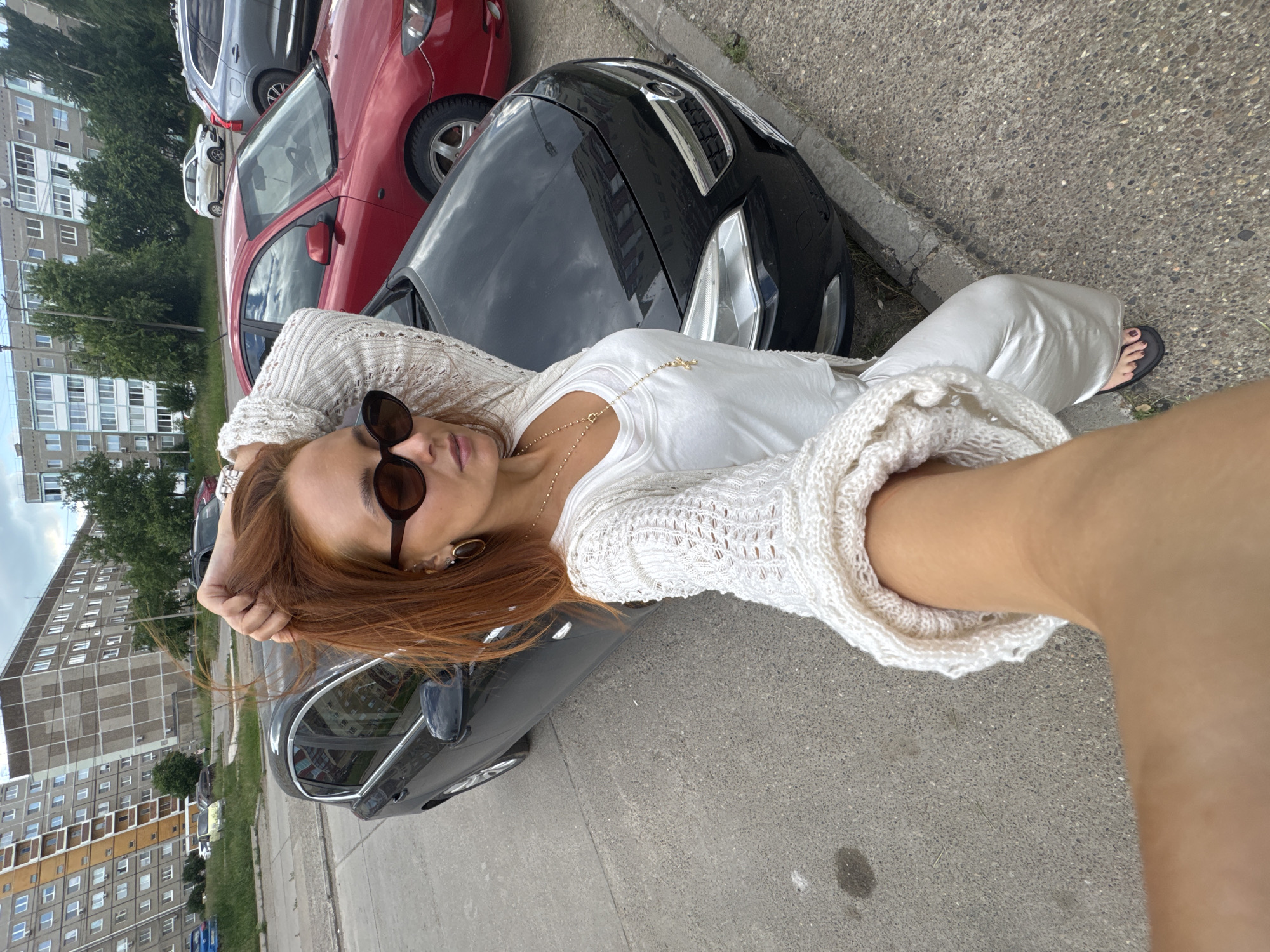 A woman with auburn hair and sunglasses is taking a selfie outdoors in front of a black car. She is wearing a white knitted cardigan over a white top and white pants. The background shows a typical residential area in Chaikovsky, Russia, with multi-story apartment buildings and parked cars. The sky is cloudy.