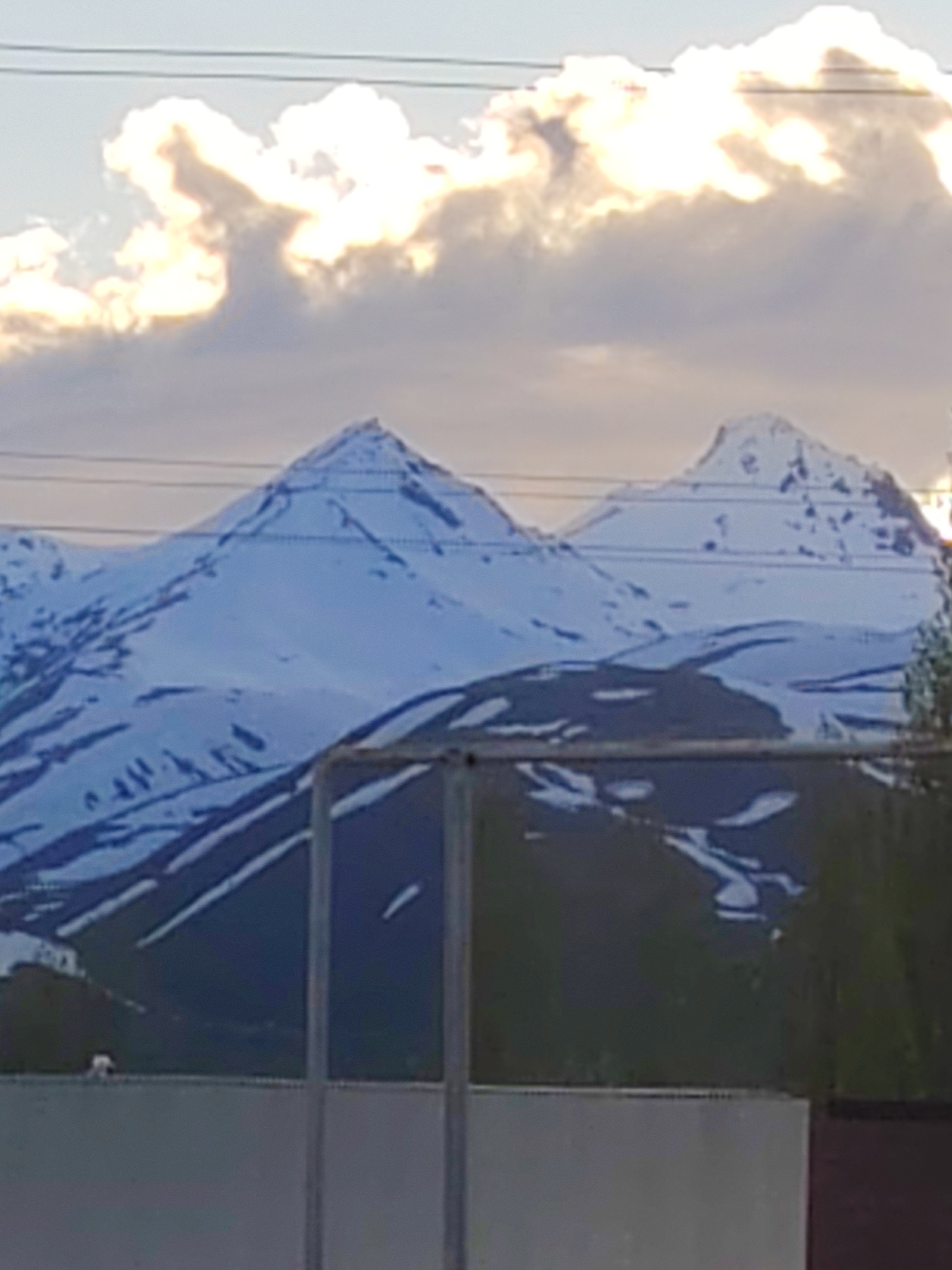 The image displays a majestic mountainous landscape, featuring two prominent snow-capped peaks under a partially cloudy sky. The mountains are extensively covered in white snow, with darker patches of rock or earth showing through, particularly on the lower slopes and in crevices. Numerous horizontal power lines or communication wires traverse the mid-ground, cutting across the face of the mountains and the sky, indicating the presence of human infrastructure.
The sky above is a mix of pale blue and muted grey clouds. Several large, bright clouds are prominently featured, glowing with a warm, golden light, suggesting either a late afternoon or early morning setting with the sun low on the horizon.
In the foreground, a dark, fence-like structure with vertical metal posts and a horizontal bar partially obscures the view of the mountains. Below this, a light-colored concrete barrier or wall stretches across the bottom of the frame. To the right, some dark green foliage, possibly trees or bushes, is visible. A small, indistinct white object rests on the concrete barrier towards the left.
The scene is tranquil, devoid of people or visible activities, and highlights the grandeur of nature juxtaposed with subtle signs of human presence. No text is visible in the image.