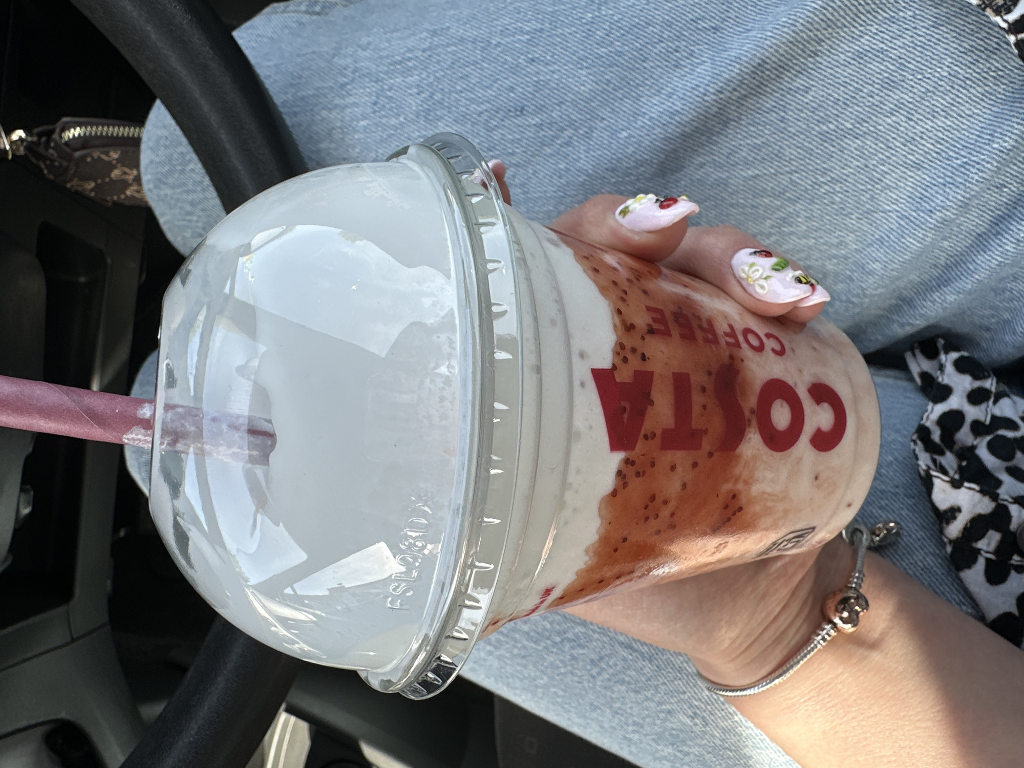 A person's hand, adorned with a silver bracelet and decorated nails featuring floral and insect designs, holds a clear plastic cup filled with a strawberry-flavored drink and topped with whipped cream. The cup has the "COSTA COFFEE" logo printed on it in red. A pink paper straw extends from the domed lid. The background shows the interior of a vehicle, including the steering wheel and dashboard, and the person's light blue denim jeans. The scene is lit by natural sunlight, suggesting it is daytime. The location is the city of Carlow, Ireland.
