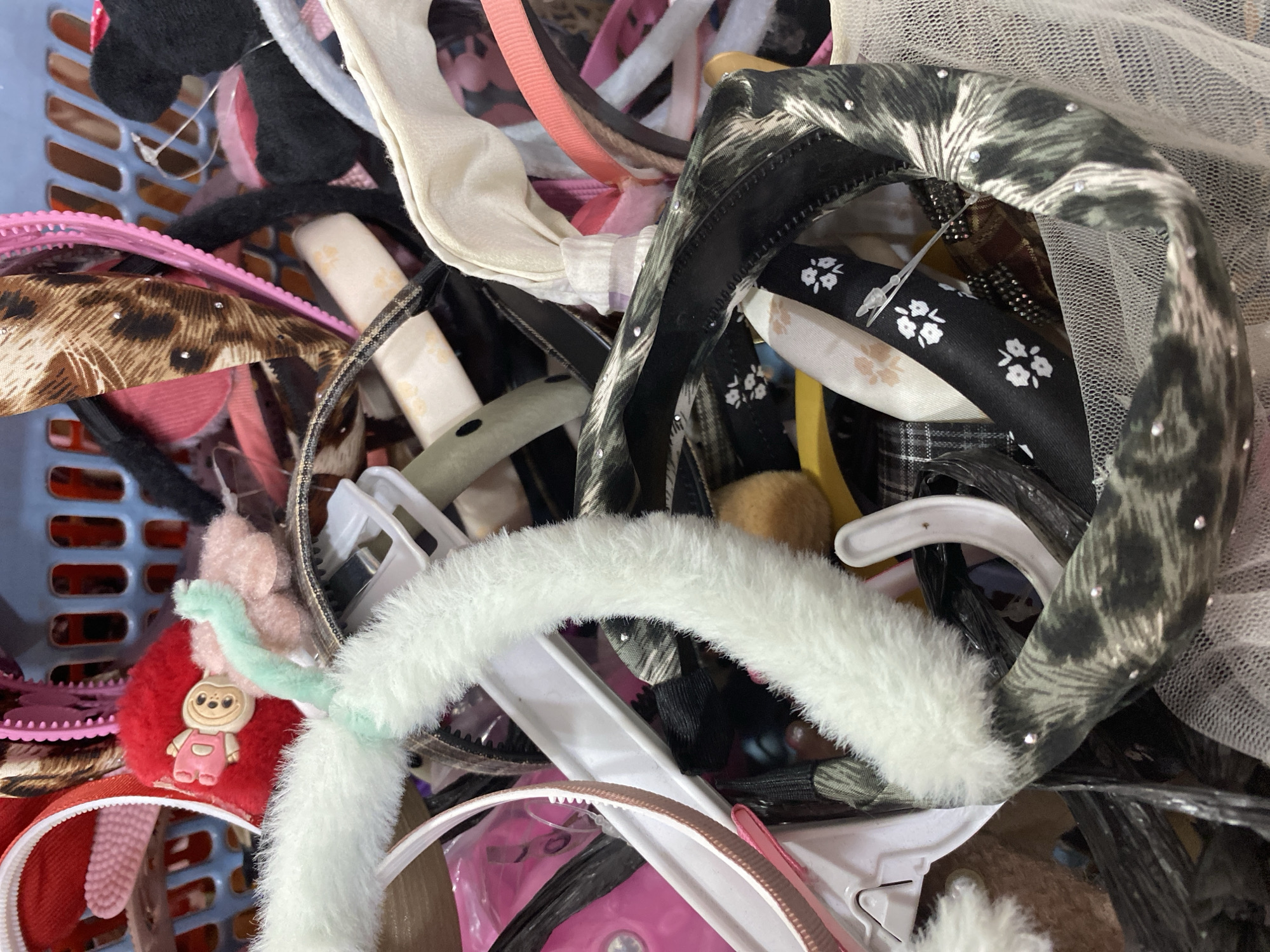 The image shows a close-up, overhead view of a tangled assortment of headbands and hair accessories in a blue plastic basket. The headbands are of various colors, patterns, and textures, including leopard print, floral patterns, fuzzy white, and plain colors. Some have embellishments like rhinestones. The basket appears to be filled to capacity, with the headbands spilling over and obscuring the sides of the basket and other potential items within. The lighting suggests an indoor setting, likely a storage area or a retail display. There are no people or visible actions depicted. The location in Lamteumen Barat, Indonesia is not evident from the image itself.