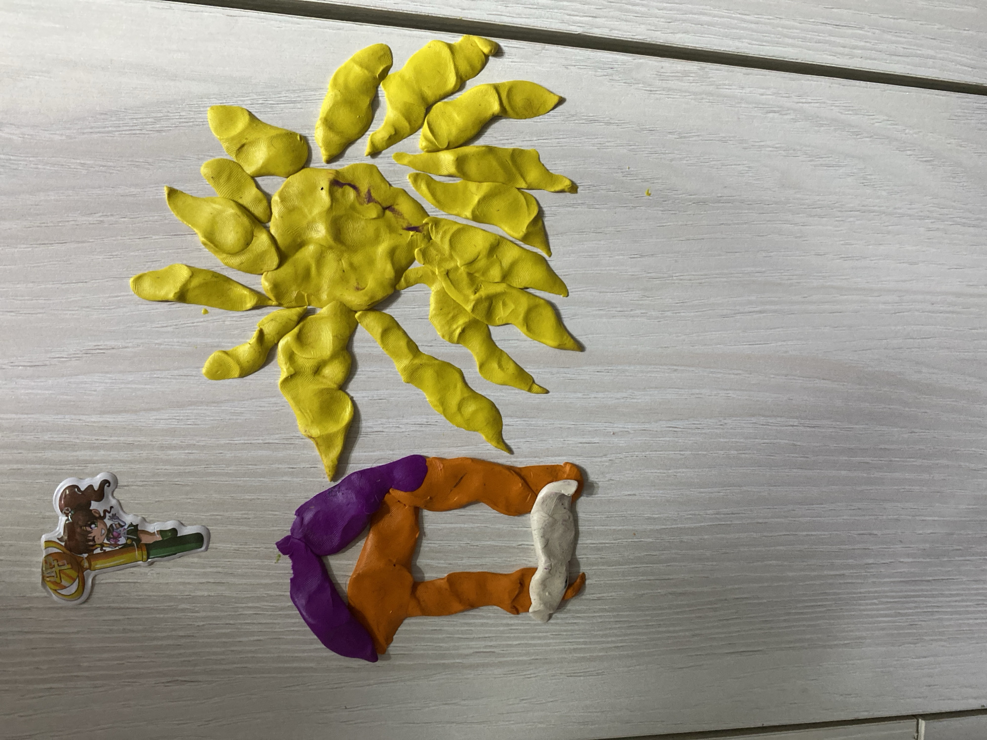 A yellow sun made of playdough is on a light grey wooden surface. Below the sun, there is a colorful rectangular shape made of purple, orange, and white playdough. To the left of the playdough shapes, there is a sticker depicting a cartoon girl with brown hair, lying on a golden object. The background appears to be a table or countertop. There is no indication of the location of Chaikovsky, Russia in the picture. There are no visible people.