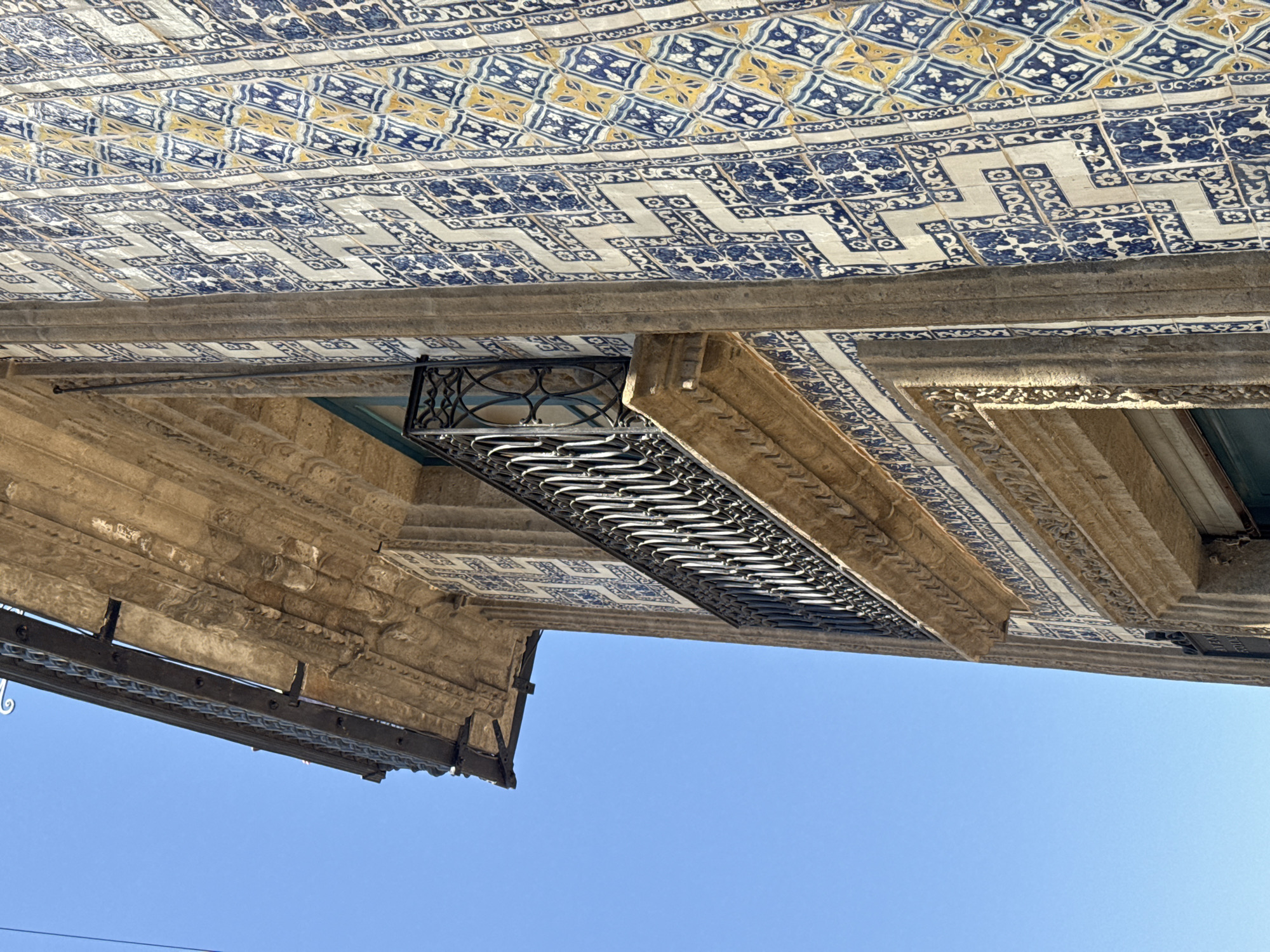 The image shows a section of a building facade in Mexico City, Mexico, viewed from below at an angle. A blue sky occupies the majority of the frame. The facade features a heavily ornamented, light-brown, stone-like structure. Decorative azulejo tilework in shades of blue, white, and yellow adorns the upper part of the facade. A dark-colored wrought iron balcony with rounded details protrudes from the building. Another lower section of a wrought iron overhang is also visible.