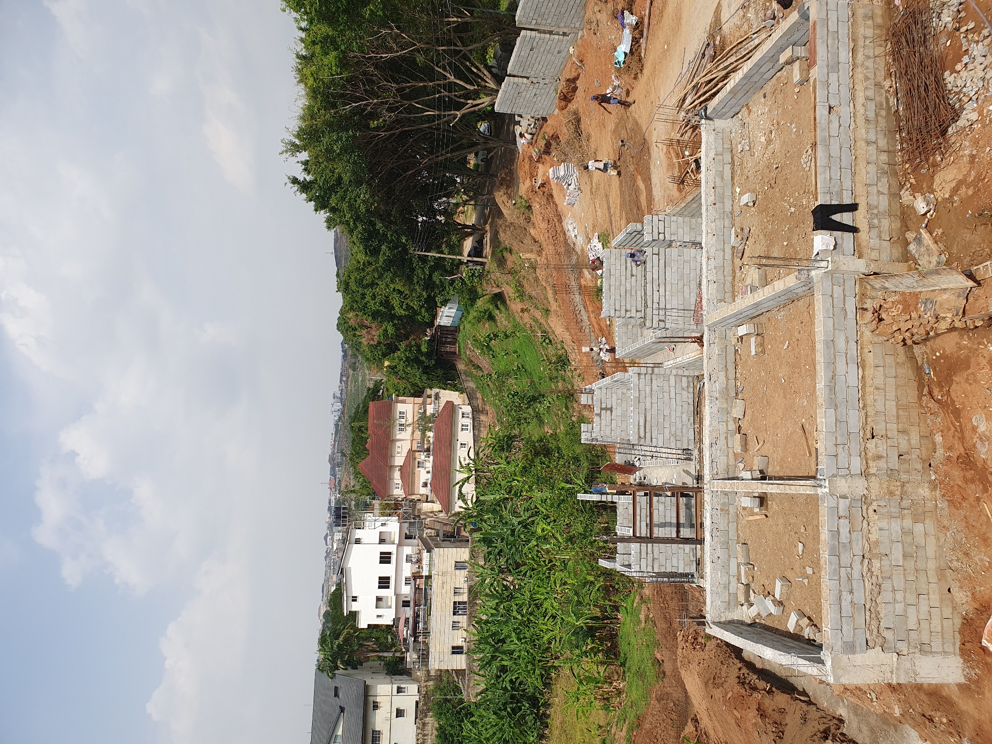 The image shows a construction site in Abuja, Nigeria. Multiple building foundations are visible, constructed from grey cinder blocks. Workers are present, moving around the site, some handling materials. Rebar, wooden planks, and piles of cinder blocks are scattered around the construction area. A backdrop of residential buildings and greenery is visible, with trees and lower-lying structures in the distance. The sky is overcast. The ground is dirt, and appears to be on a slope.