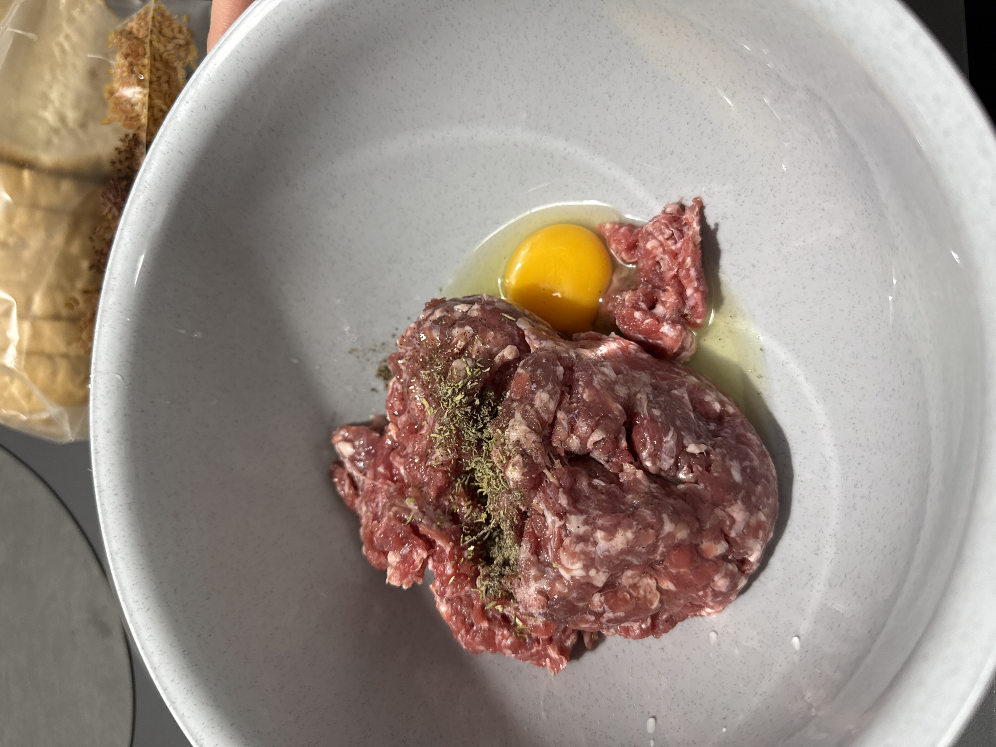 A grey bowl contains raw ground meat seasoned with dried herbs and a raw egg. A package of bread is visible to the left of the bowl. The location and city are not identifiable from the image.