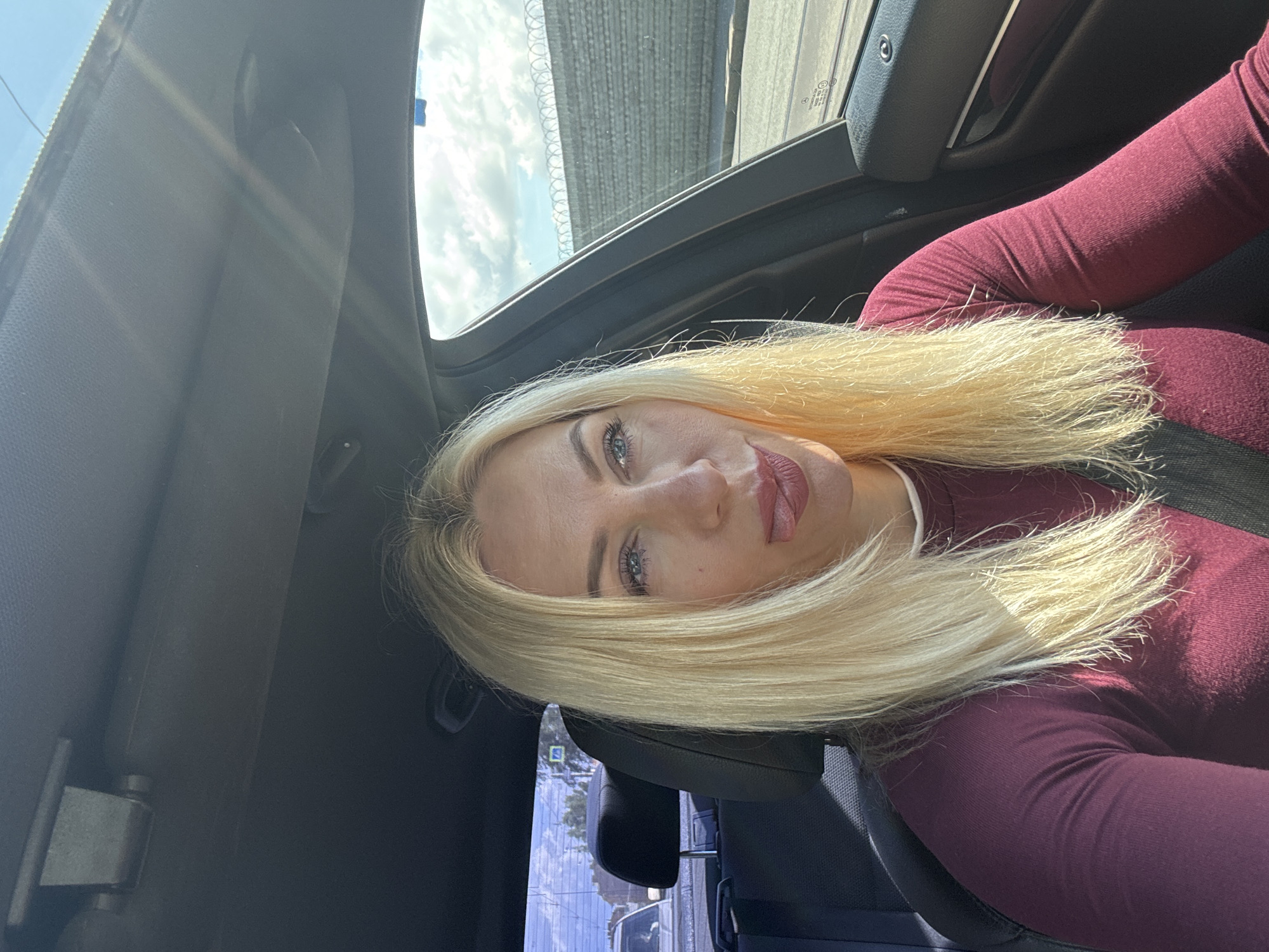 A woman with blonde hair is sitting in the driver's seat of a car, sticking her tongue out. She is wearing a maroon long-sleeved shirt and a black seatbelt. The car is dark-colored on the interior. Outside the car, there is a cloudy sky and a stone wall with barbed wire on top. A street is visible in the background, along with other cars and some buildings. The scene appears to be in an urban setting, possibly Saint Petersburg, Russia, given the architectural style of the wall and the presence of street signs. The lighting suggests it is daytime.