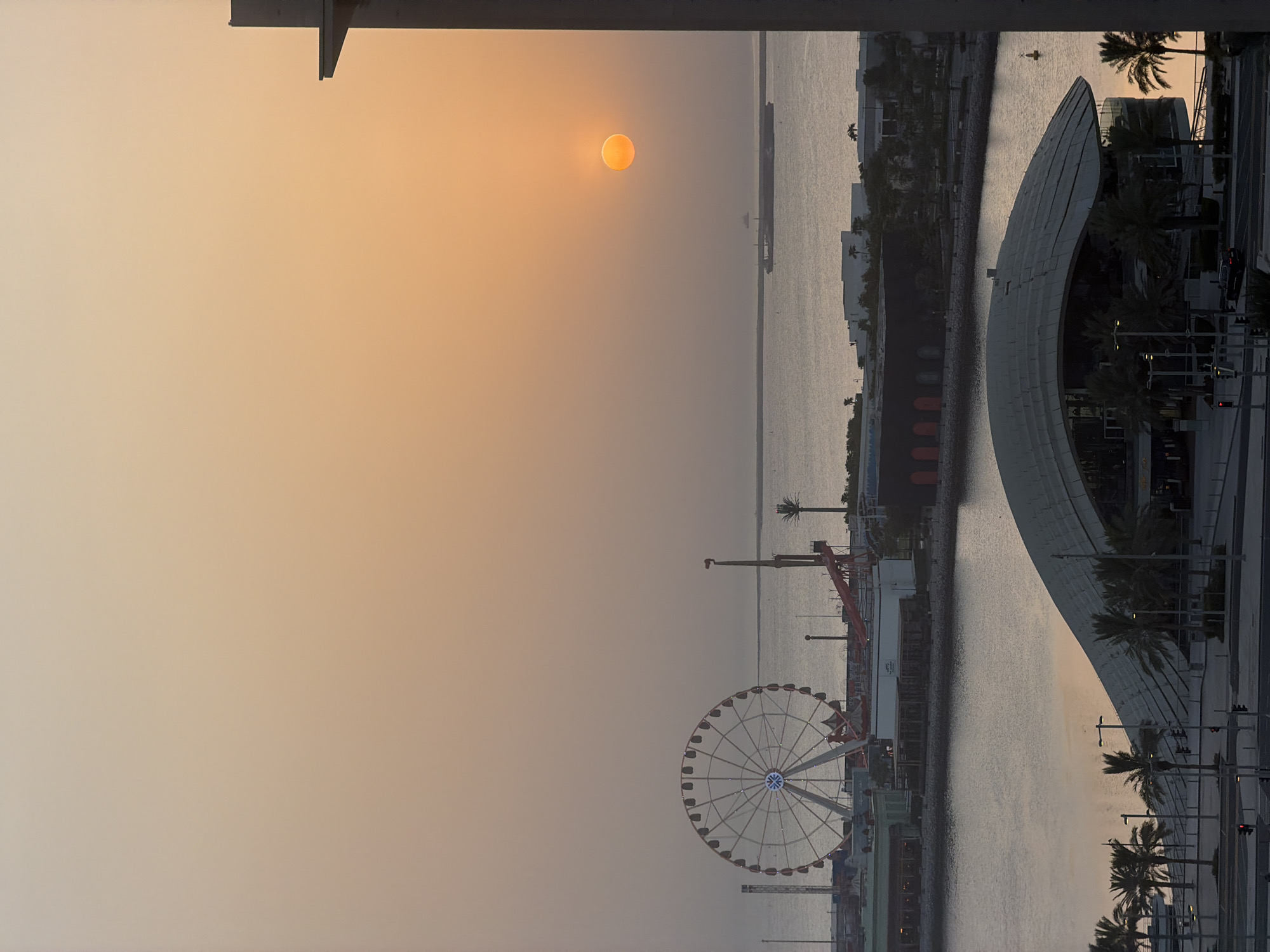 This is an aerial, vertical, high-angle, full shot of the waterfront in Lusail, Qatar, at sunset. The sky is a hazy orange and yellow gradient. The sun is a bright orange orb positioned in the upper right quadrant of the image. A large Ferris wheel, lit with white and blue lights, is located on the left side of the frame, near the waterfront. Several buildings with dark facades and red accents are visible behind the Ferris wheel. In the foreground, a large, modern building with a curved, white roof is prominent, surrounded by palm trees. The water of the bay is calm and reflects the colors of the sky. A large cargo ship is visible in the distance on the water. A building structure, possibly a balcony or ledge, is in the far right foreground. The scene depicts a tranquil, coastal urban environment during twilight.