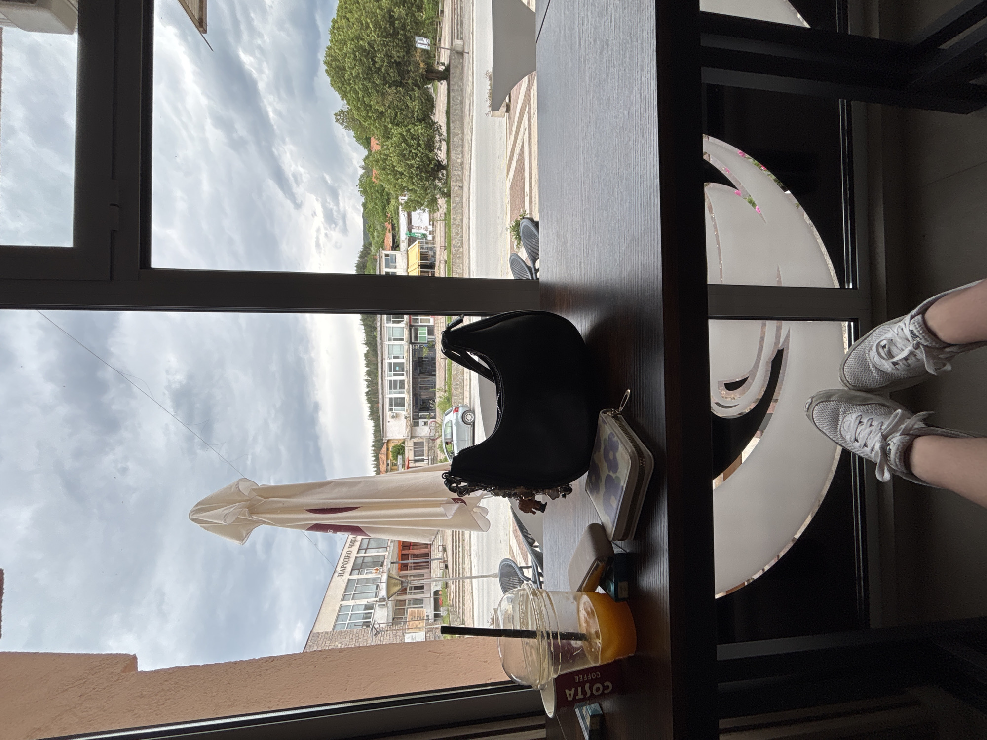 From an indoor setting, a view through a window frame reveals an outdoor cityscape. In the foreground, on a table, there's a cup of Costa Coffee with a straw, next to a black handbag and a wallet. The exterior view encompasses buildings, streets, and trees under a cloudy sky. An open umbrella can be seen in the view. A person is positioned within the scene, as evidenced by a pair of legs and sneakers visible in the lower right corner. The location is identified as Garmen, Bulgaria.