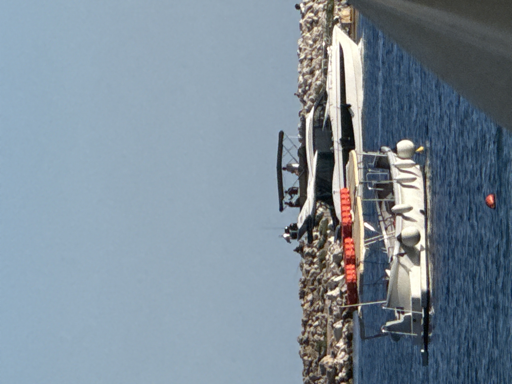 The image displays a daytime maritime scene in Budva, Montenegro, under a clear light blue sky. A large, white and black motor yacht, equipped with a flybridge and an overhead canopy, traverses the mid-ground from left to right, producing a minimal stern wake. Four individuals are visible on the yacht's flybridge, and at least one person is discernible on its main deck. Behind the yacht, a rocky breakwater or shoreline stretches horizontally, populated by several indistinct human figures. In the foreground, a smaller, white open motorboat with a light-colored canopy remains stationary or drifts. This smaller vessel features multiple white cylindrical fenders along its port side and a storage rack at the stern containing several orange life jackets or buoys. A single, small spherical orange buoy floats in the water to the immediate right of the smaller boat. A dark gray, out-of-focus structure partially intrudes into the lower right corner of the frame.