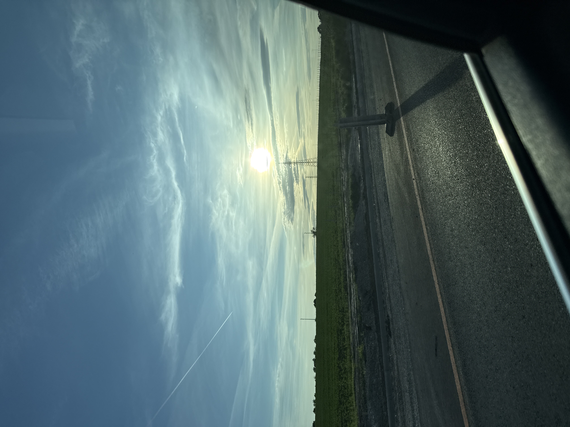 A roadside view from inside a vehicle. The scene captures a bright sun illuminating a field with power lines. A portion of the road with a dividing line is visible to the right, with the interior of the vehicle visible on the far right and top. A contrail bisects the upper left blue sky. The location of the photo is unknown. There are no people visible.