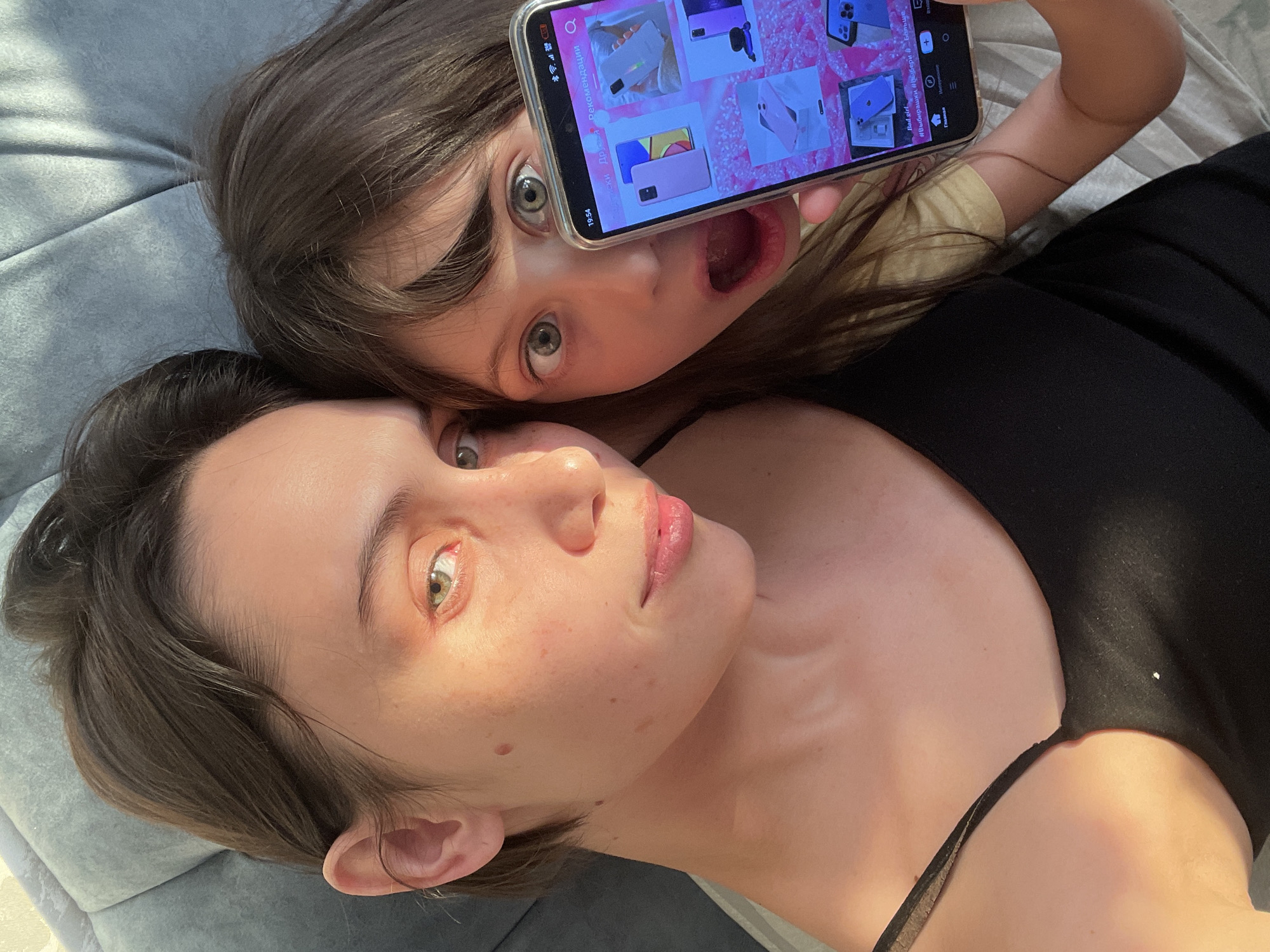 A selfie shows a woman and a child lying on what appears to be a gray upholstered surface. The woman has short brown hair and fair skin, while the child has longer brown hair, light eyes, and an open mouth. The child is holding a smartphone up to their face, obscuring part of it, and the phone screen displays multiple images of phone cases. The woman is wearing a black top with thin straps. The location is not discernible as being in Izhevsk, Russia.