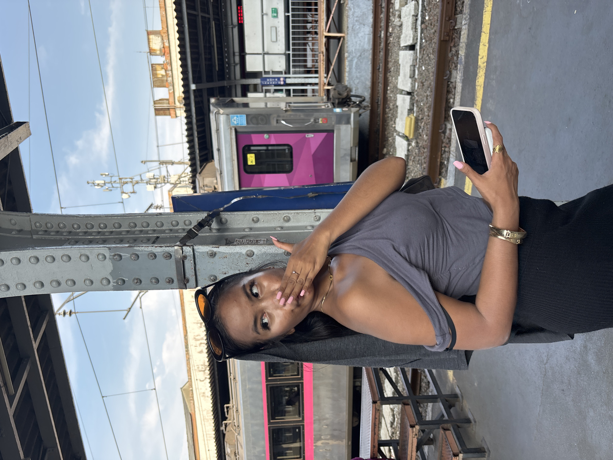 A woman stands at a train station in West Jakarta, Indonesia. She holds a smartphone in her right hand. She is wearing a gray tank top, a black skirt, and a gold bracelet. Sunglasses are placed on top of her head. A purple and silver train is in the background. The train station platform is made of concrete.