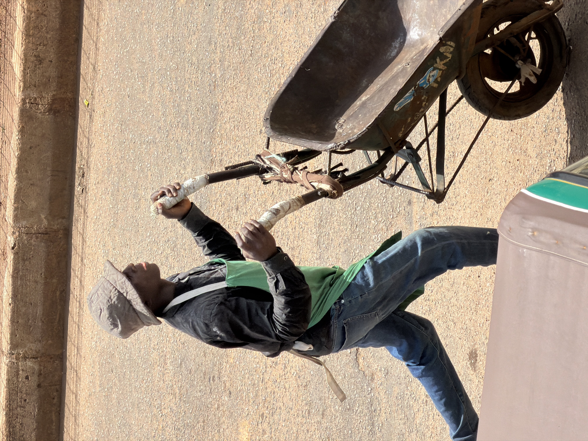 A Black man wearing a bucket hat, black shirt, green apron, and blue jeans pushes a rusty wheelbarrow on a paved road in Bukuru, Nigeria. The wheelbarrow has a metal basin and a single wheel with a worn tire. The man is holding the handles of the wheelbarrow, and his body is angled as he walks. The road is a light brown color and appears to be made of asphalt or a similar material. In the background, there is a concrete barrier or curb. The scene is illuminated by bright sunlight, casting shadows. The image is taken from a high angle, looking down on the man and the wheelbarrow. A portion of another vehicle, possibly a tricycle or motorcycle sidecar, is visible in the lower left corner of the frame. The wheelbarrow has some graffiti or markings on its side, including what appears to be the word "AKAI."