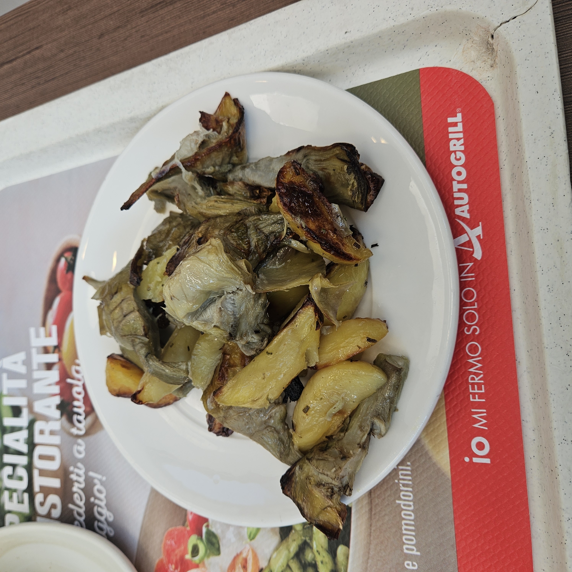 A close-up shot shows a white plate of roasted artichoke hearts and potato wedges on a white tray. The plate is slightly off-center to the right. The background features a blurred image of restaurant specials and a red paper napkin with the text "io MI FERMO SOLO IN AUTOGRILL." The overall impression is that of a meal being served at an Autogrill service station along the A14 highway, which runs through the city of Riccione, Italy.