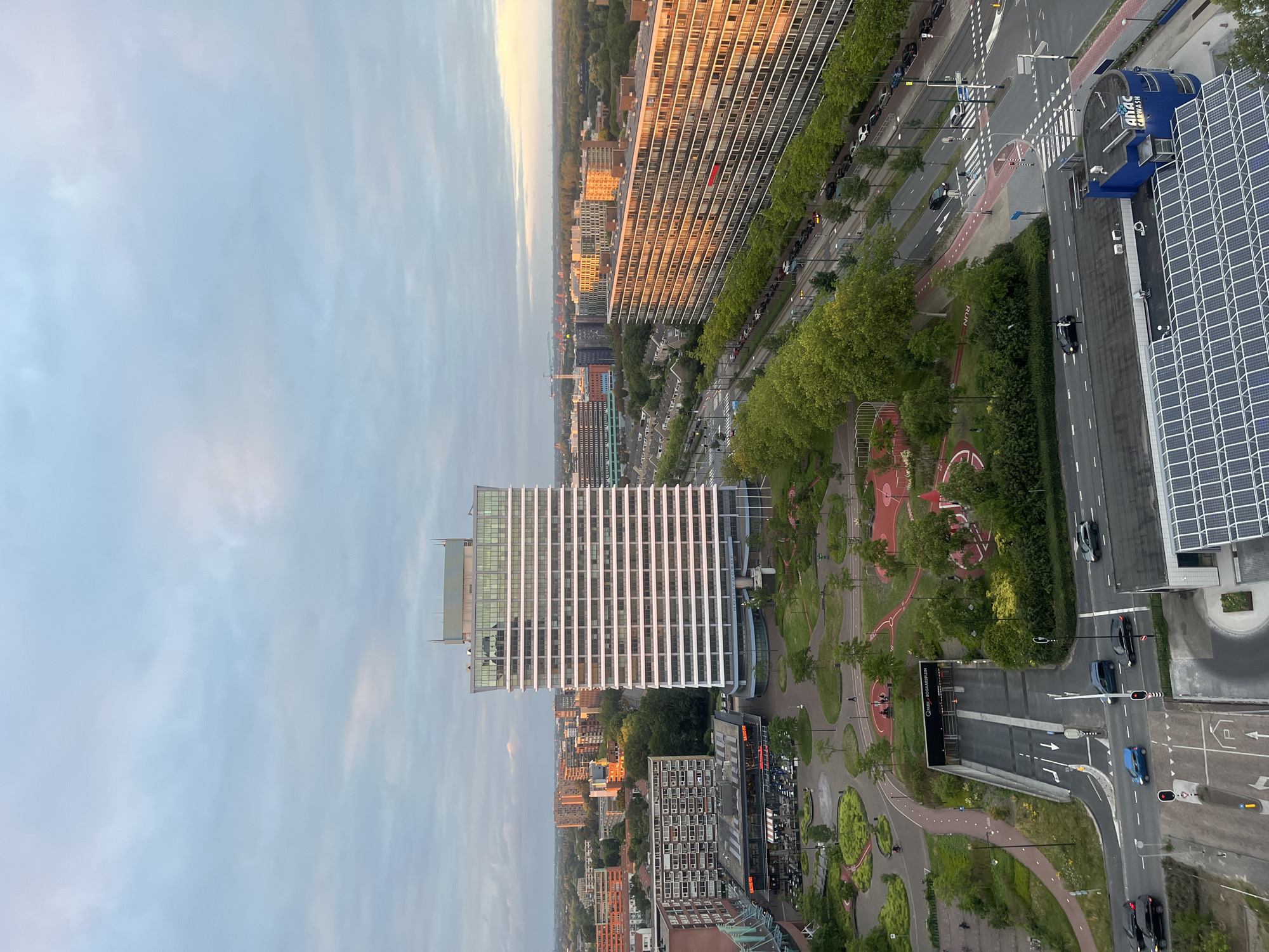 This is an aerial, high-angle, full shot of a cityscape. The sky is filled with soft, pale blue clouds. In the center of the image, a tall, modern white building with many horizontal windows dominates the view. To the right of this building, a long, lower, apartment complex with balconies is visible, reflecting the warm orange and yellow light of the setting sun. In the foreground, a multi-lane road is visible with several cars driving on it. To the left, an underground parking garage entrance, labeled "Q-PARK BOOGAARDPLEIN," is situated, with traffic lights and pedestrian crossings nearby. A green park area with red running tracks and trees is also present in the midground. In the background, a dense urban landscape with various buildings and trees stretches to the horizon, with some industrial structures visible in the far distance. The lighting suggests it is late afternoon or early evening.