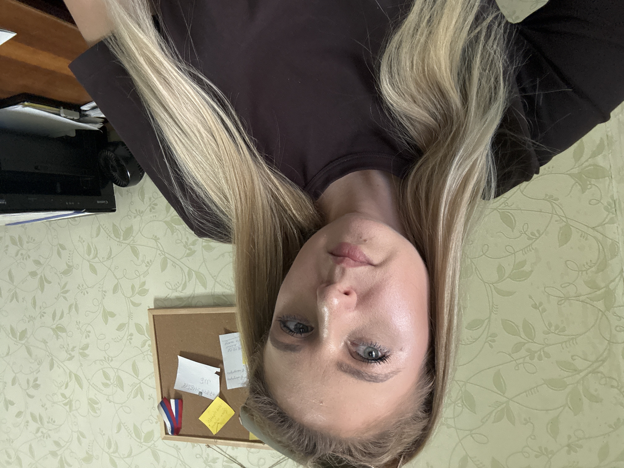 This is a close-up, upside-down selfie of a young Caucasian woman with long, blonde hair. She is wearing a dark, long-sleeved top. Her face is the primary focus, with her light blue eyes looking towards the camera. Her skin appears smooth and well-lit, with some subtle makeup, including mascara and lipstick.
Behind her, the background consists of a wall with light green, floral wallpaper. To the left of the frame, a wooden desk or shelf is visible, with some papers and a black electronic device. A small fan is attached to the side of the black device. In the lower part of the image, a corkboard is attached to the wallpaper, with several sticky notes and a small ribbon with the colors of the Russian flag.
The lighting suggests an indoor setting, possibly during the daytime, as there are no strong shadows indicating artificial light sources. The woman's expression appears neutral or slightly pensive. There is visible text on the sticky notes on the corkboard, including what appears to be a phone number and some Cyrillic script. The overall mood of the image is casual and personal, typical of a selfie.
