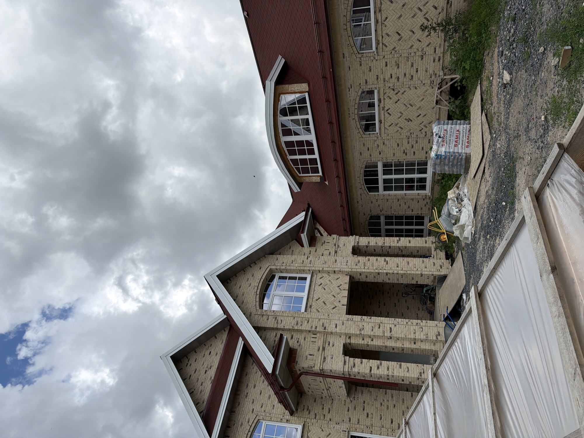 The image shows a multi-story house under construction in Kamyshevakha, Russia. The house features a light-colored brick exterior with a dark red tiled roof. Several windows are visible, including arched windows on the upper floors. Construction materials, such as bags of cement and plywood sheets, are scattered around the property. A covered structure, possibly a greenhouse, is in the foreground. The sky is overcast with gray clouds.