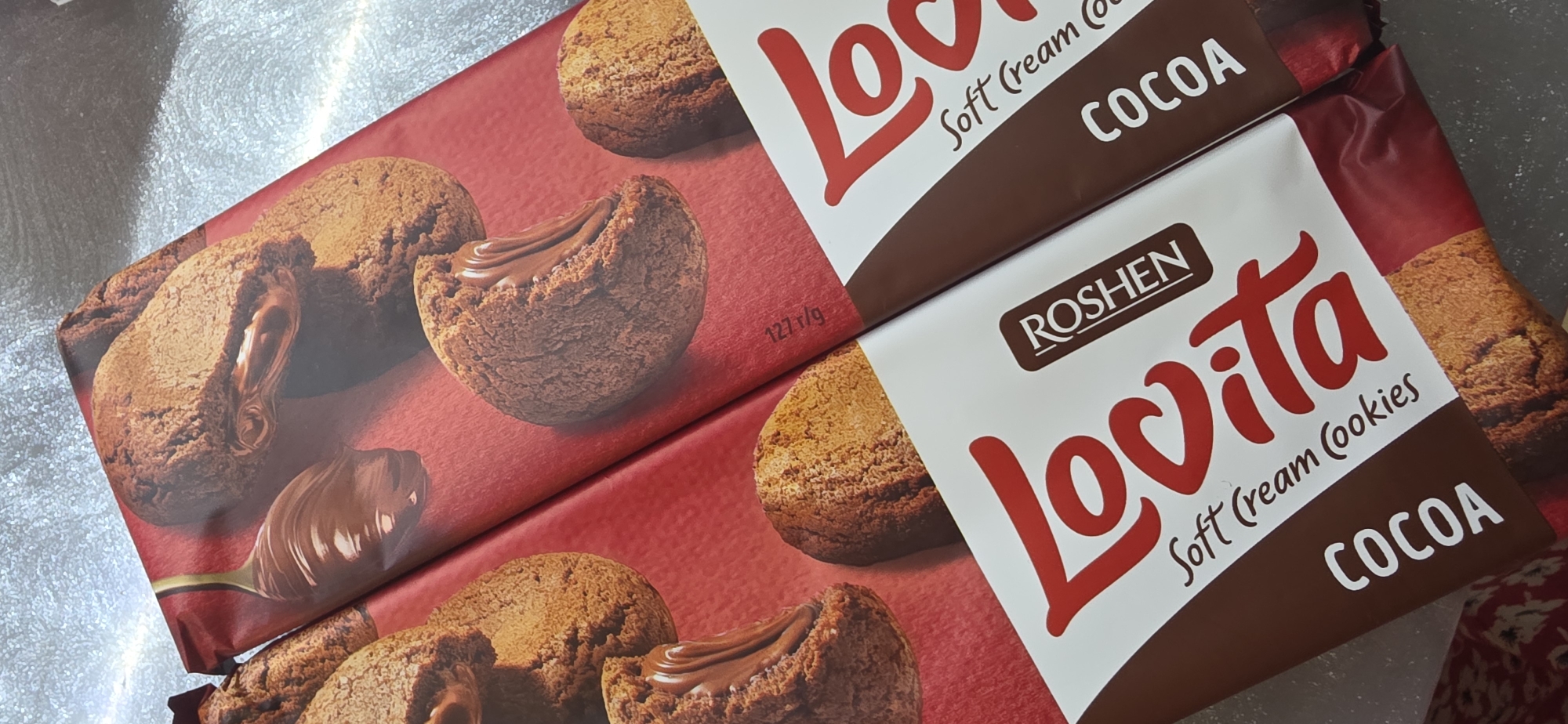 Two packages of cookies are displayed diagonally on a metallic surface. The packages have a red background with images of chocolate-filled cookies. The branding on the packages reads "Lovita" in large red letters, with "ROSHEW" and "Soft Cream Cookies COCOA" in smaller text. The cookies themselves appear to be round, dark brown, and have a textured surface. Some of the cookies are broken open, revealing a creamy chocolate filling. The metallic surface has a brushed texture and reflects light, suggesting it might be a countertop or table. There are no people visible in the image. The location of the image is not specified in the visual content, and therefore cannot be determined to be in the city of Sumgayit, Azerbaijan.