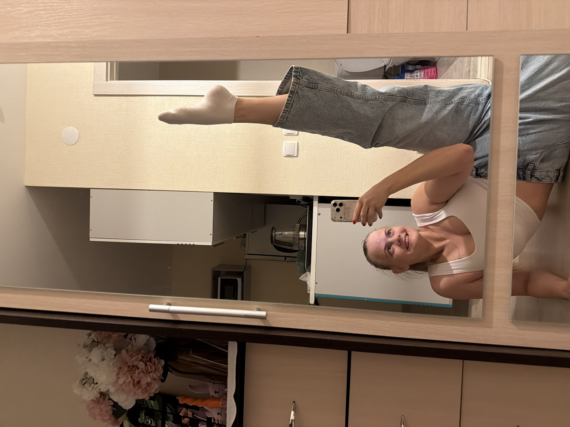 A person in a white top and light blue jeans is photographed in a mirror in Izhevsk, Russia. The person's right leg is extended, wearing a white sock. They are holding a phone and smiling at their reflection. The background reveals a kitchen setting with cabinets and appliances. A bouquet of flowers is also visible in the lower left corner.