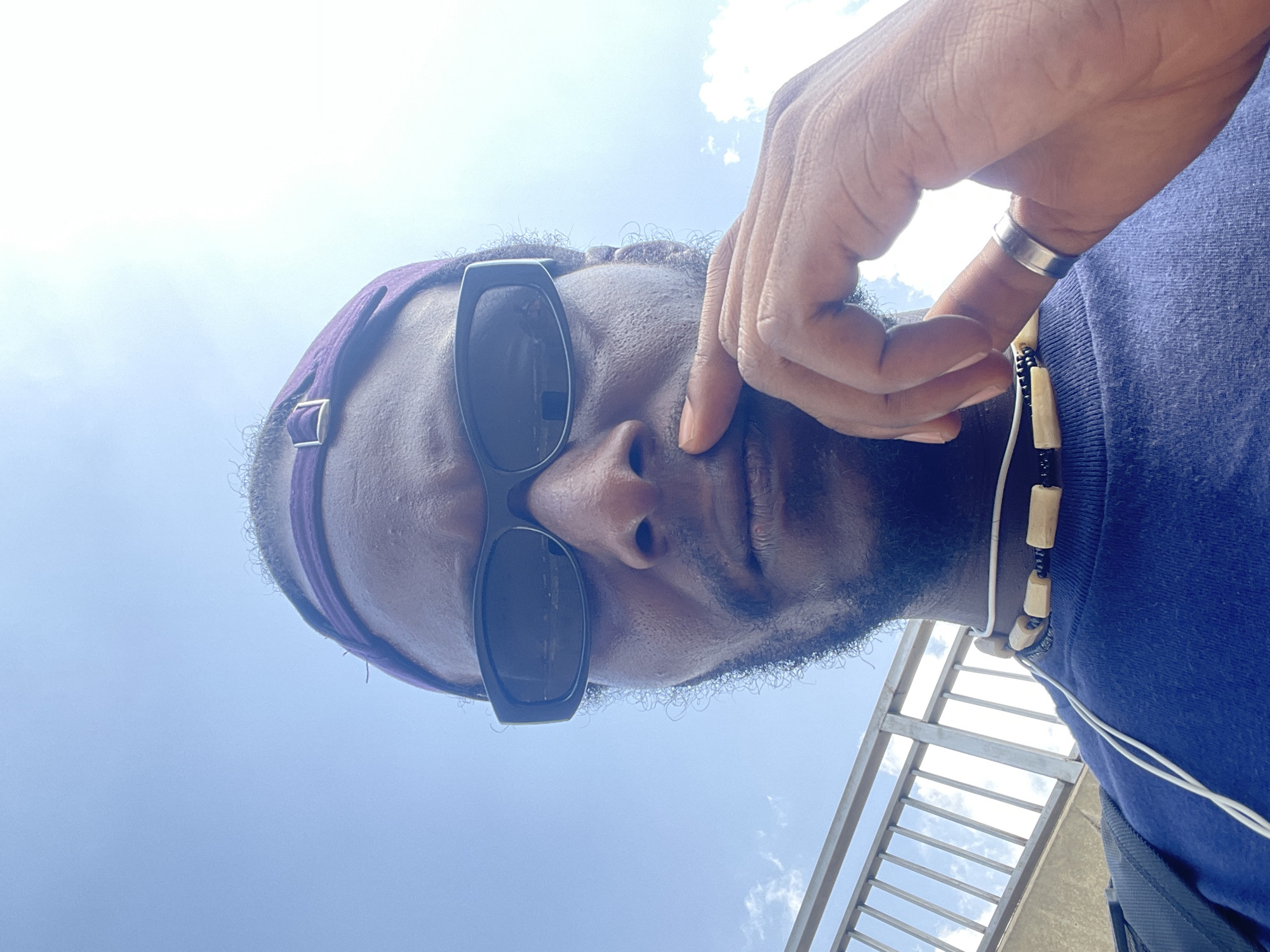 The image shows a close-up, low-angle view of a Black man wearing sunglasses and a purple headband. His hand is raised to his face. He is wearing a blue shirt and a beaded necklace. The background reveals a bright, cloudy sky and a portion of a metal railing or staircase. The scene is likely outdoors. There is no information about the location in the city of Dutsen Alhaji, Nigeria.