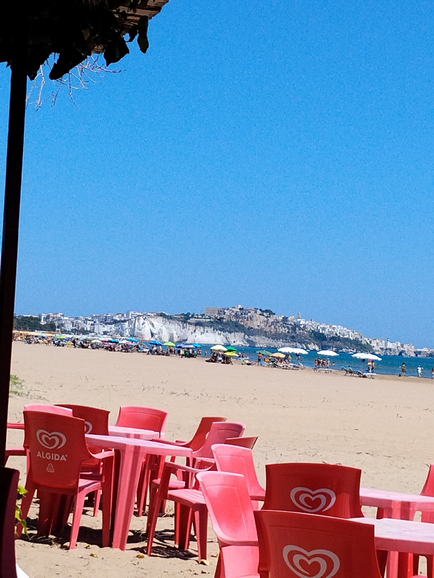 The image shows a sunny beach scene in Vieste, Italy, with the town of Vieste visible in the background. In the foreground, there are several pink plastic tables and chairs set up on the sand, some with the "Algida" ice cream logo visible on the chairs. Beyond the tables and chairs, a sandy beach is populated with beachgoers, colorful umbrellas, and lounge chairs. The sea is visible in the middle ground, with a light blue sky dominating the upper portion of the image. The coastline of Vieste, with its white cliffs and buildings, forms a picturesque backdrop.