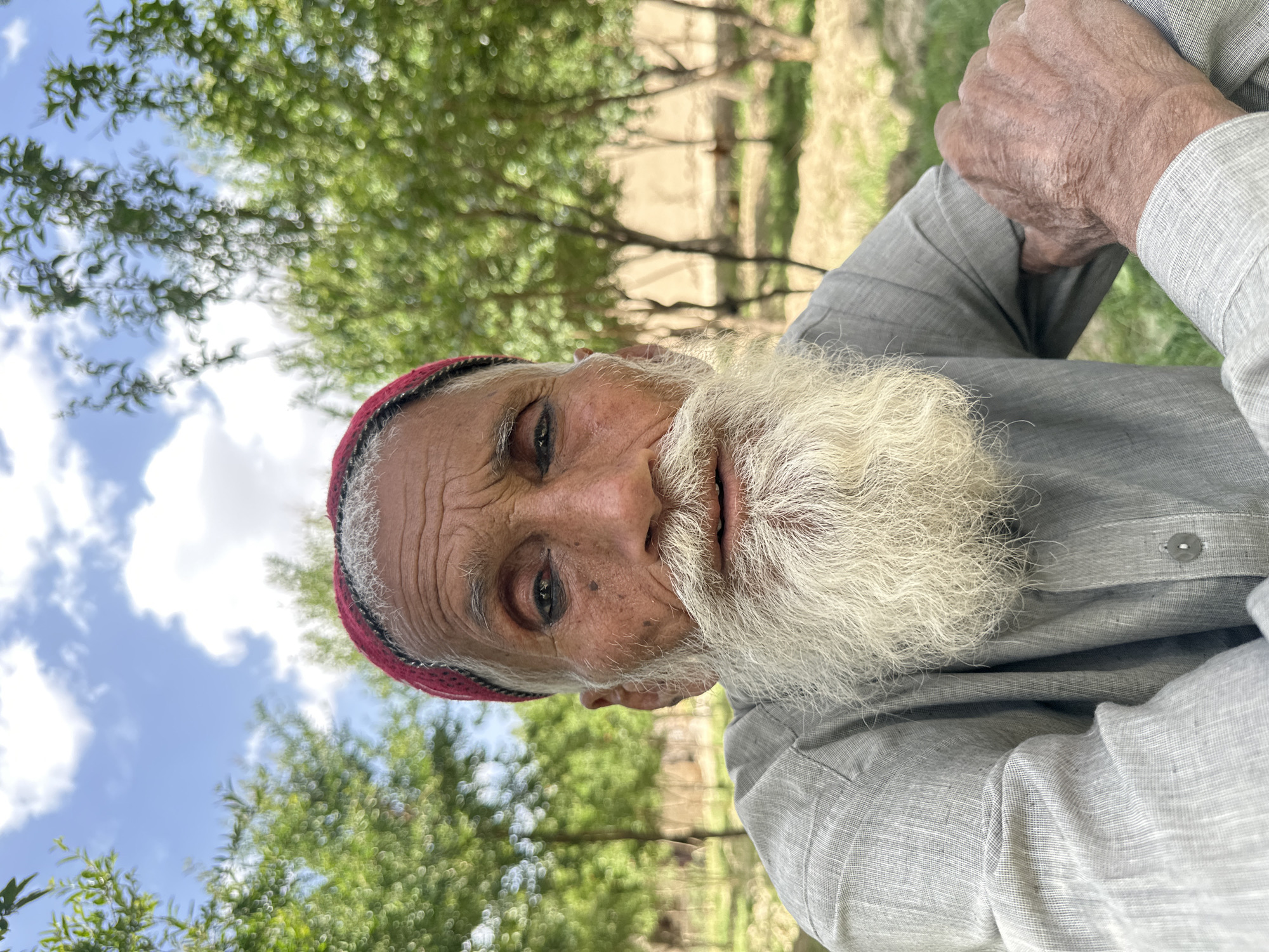 The image depicts an elderly man with a long white beard and a red cap. He is wearing a light gray shirt. He appears to be outdoors, with trees and a cloudy sky visible in the background. His hands are clasped together. There is no definitive information in the image to pinpoint the exact location in Quetta City Tehsil, Pakistan, though the environment suggests a somewhat rural area.
